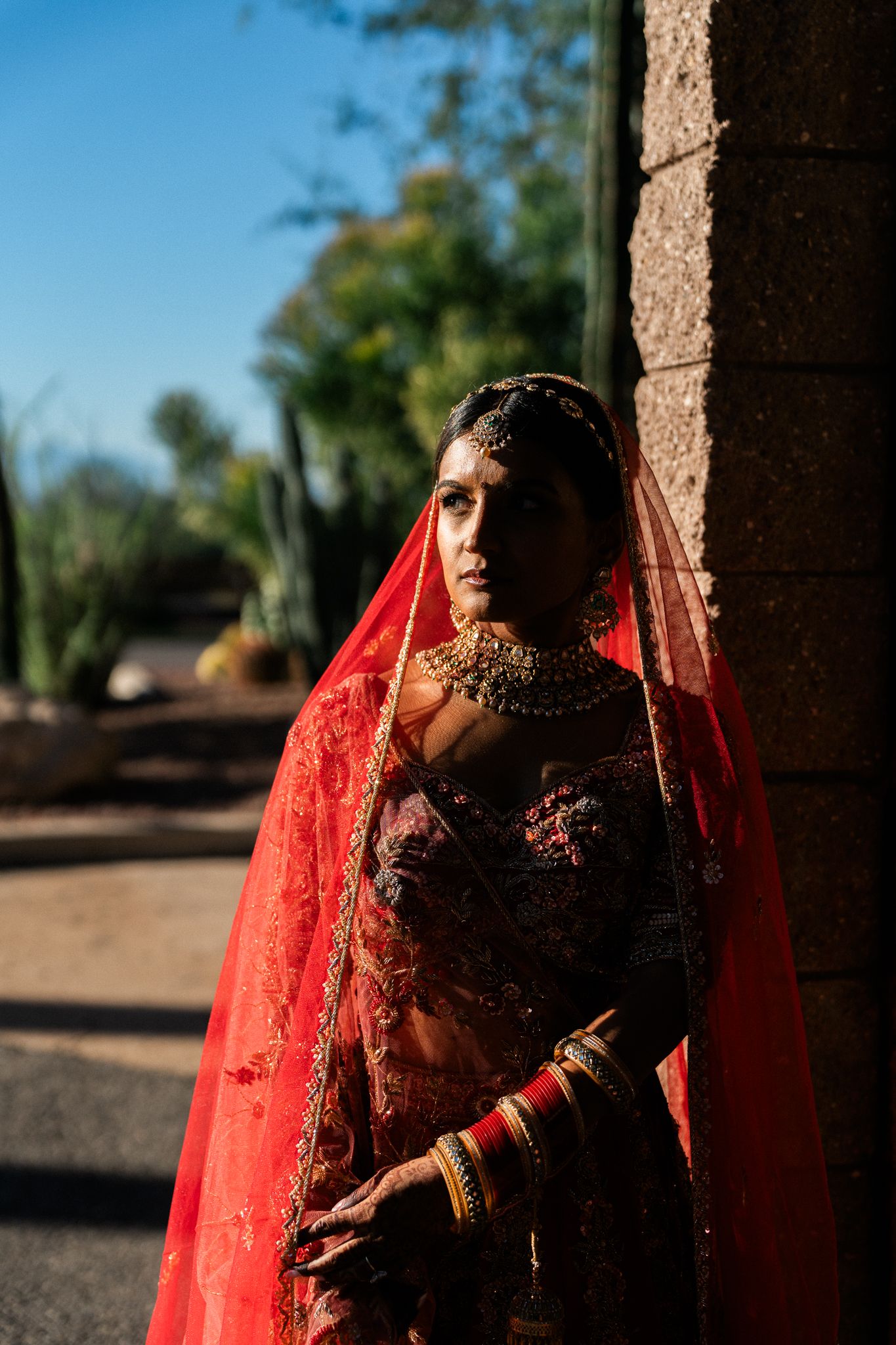 Traditional bride in red attire and veil poses dramatically in shadowed archway.