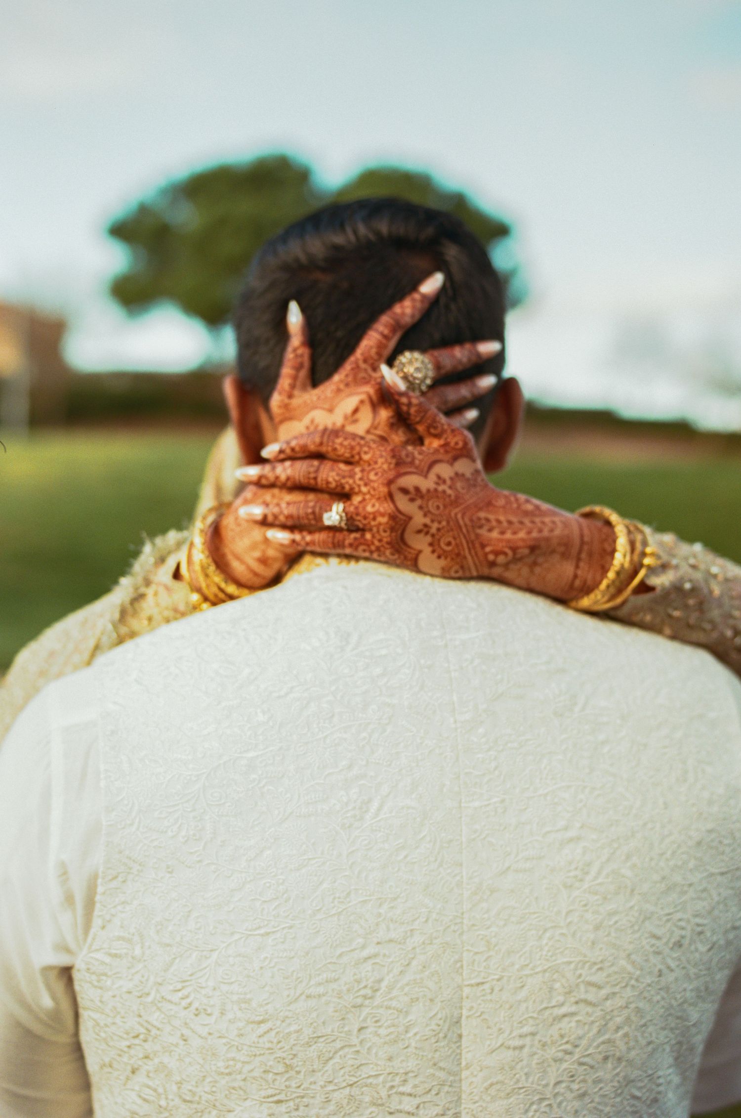 Close-up detail of traditional henna adorned hands with gold bangles against a white outfit in an outdoor setting.
