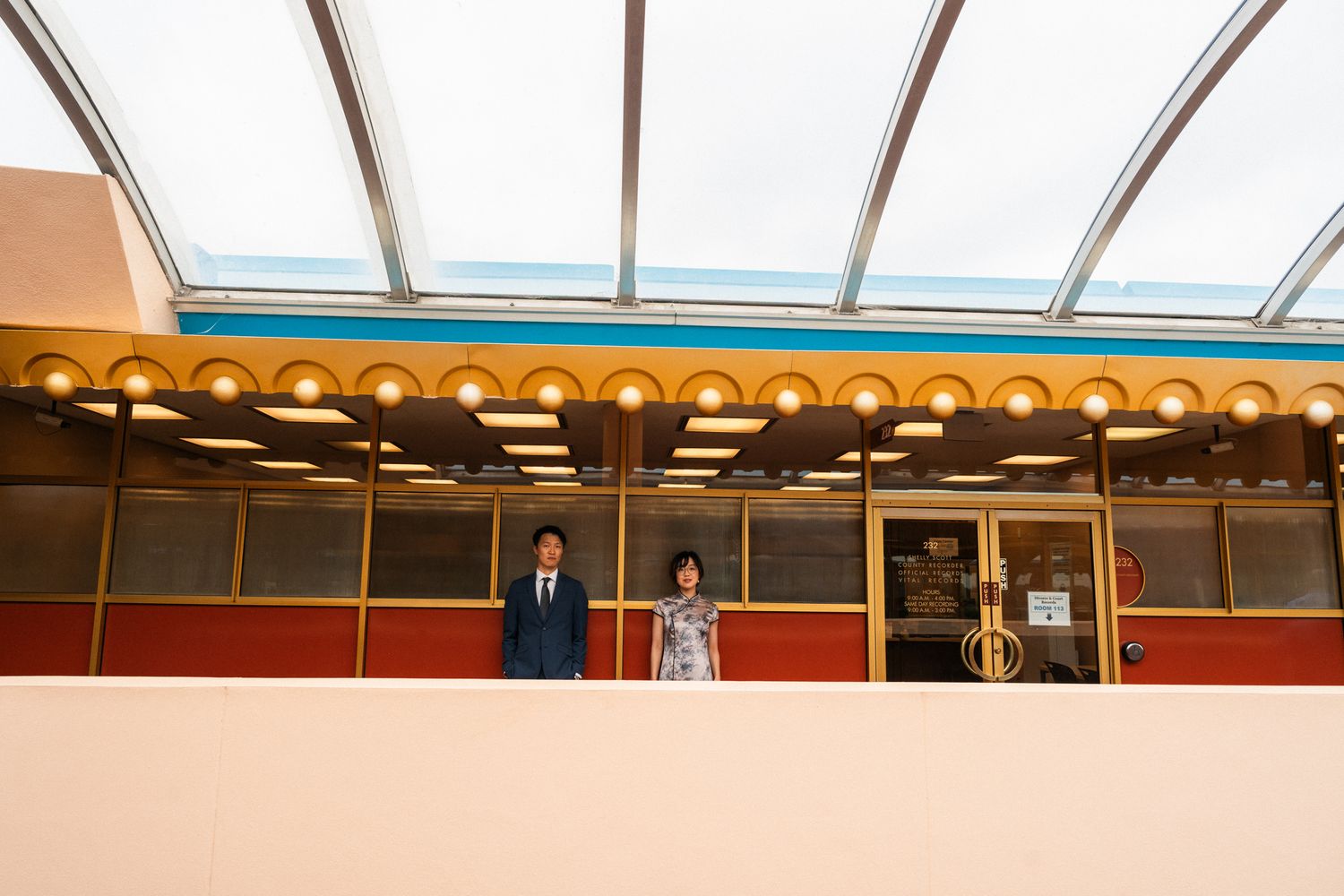 Couple standing beneath decorative arcade with glass ceiling, colorful facade, and warm lighting at a venue.