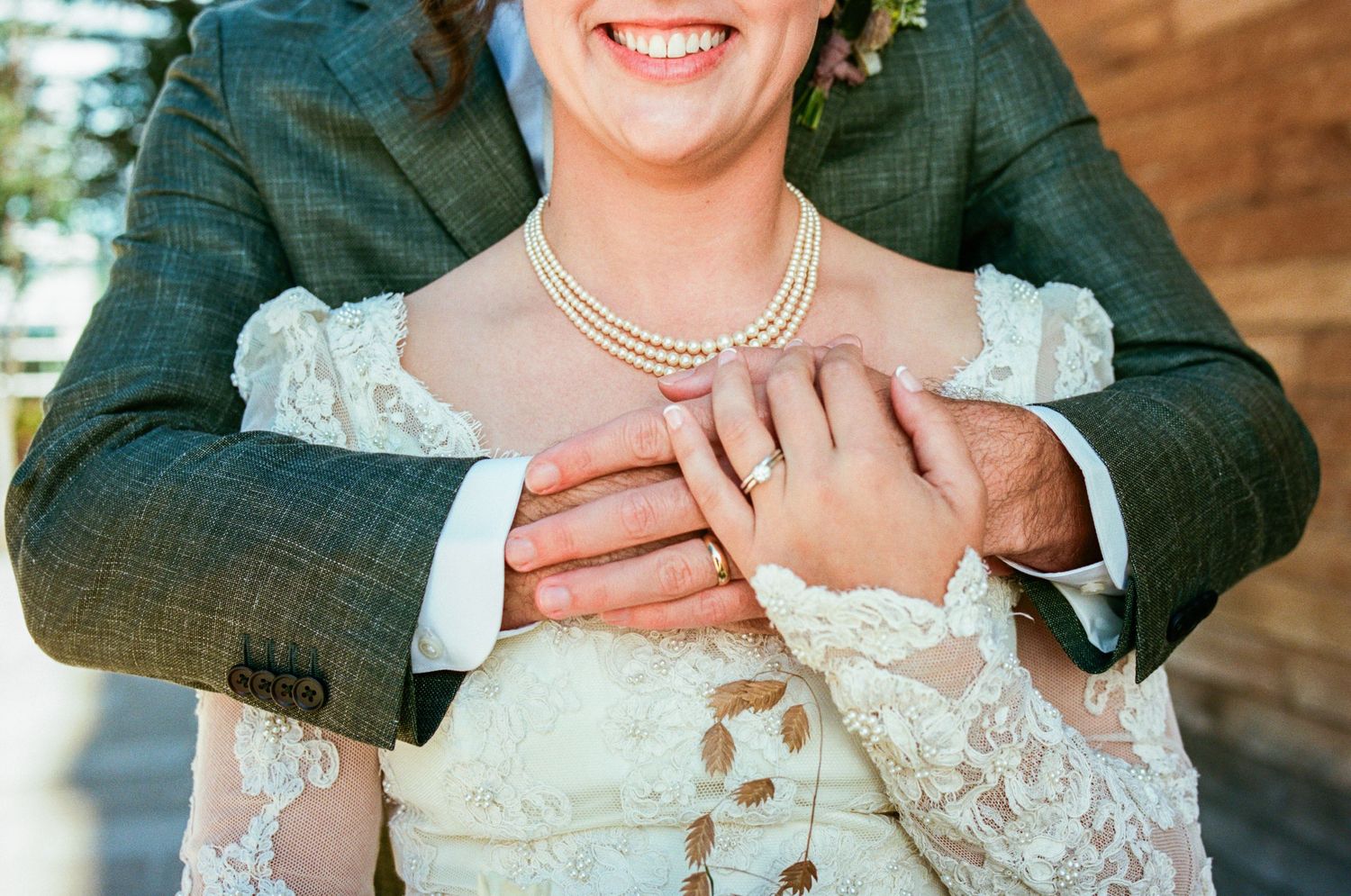 Close-up of pearl necklace and lace wedding dress details with hands showing ring.