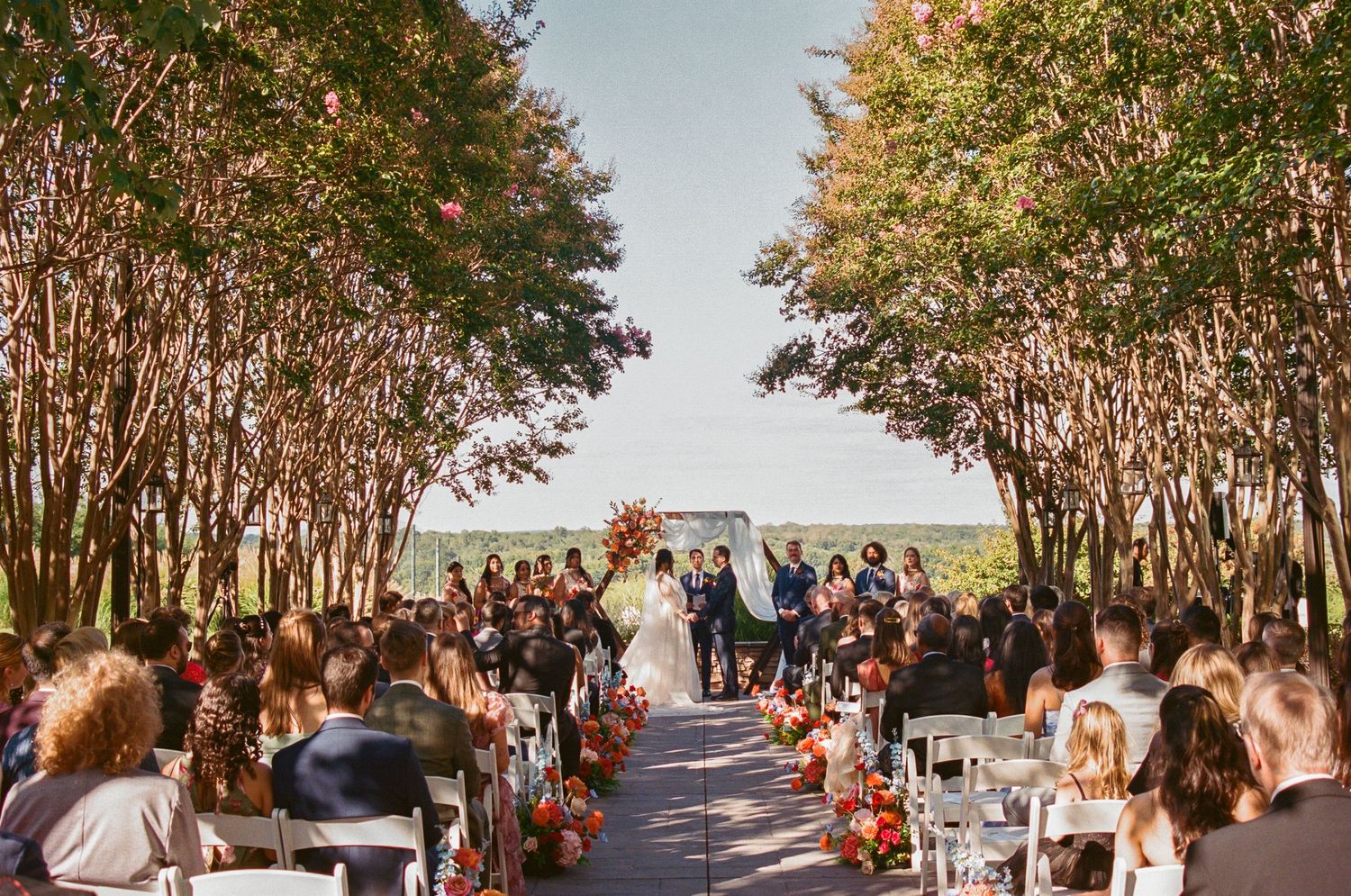 Tree-lined ceremony venue with guests seated in rows under autumn foliage.