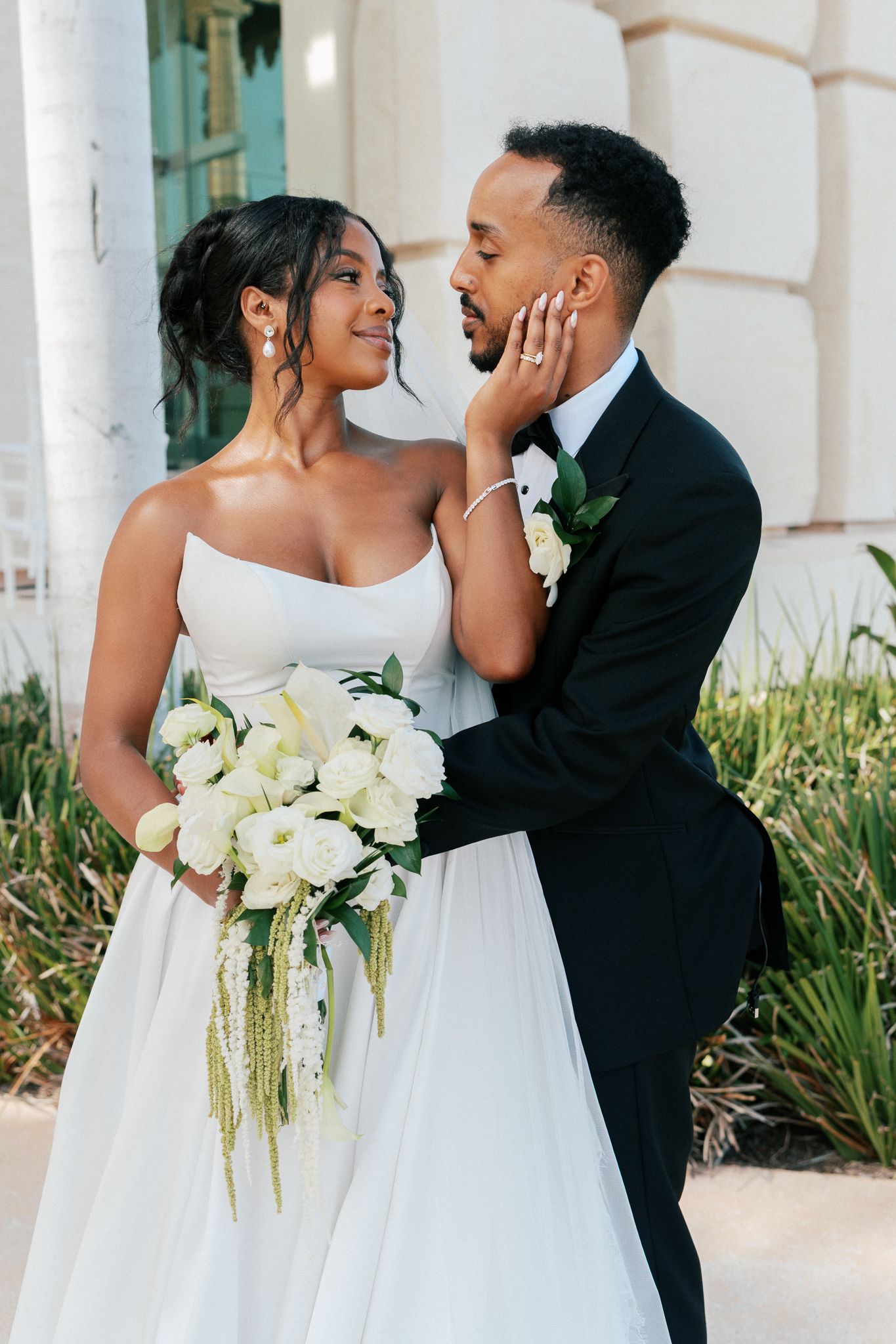 A bride and groom share a tender moment, she holds white roses bouquet while gently touching his face in wedding attire.