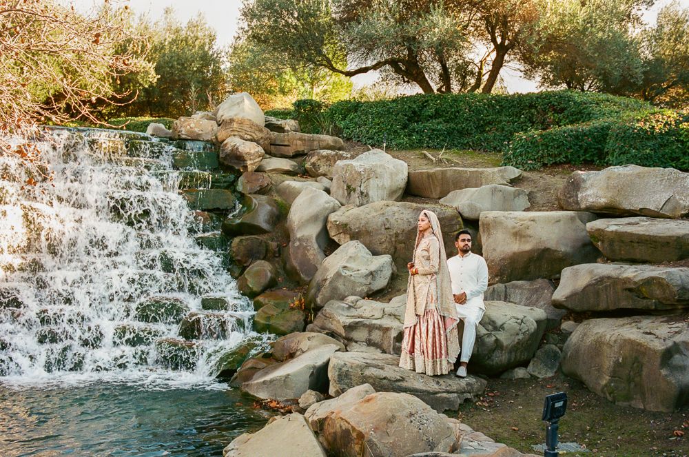 Couple in traditional South Asian attire posing by a rocky waterfall surrounded by lush greenery.