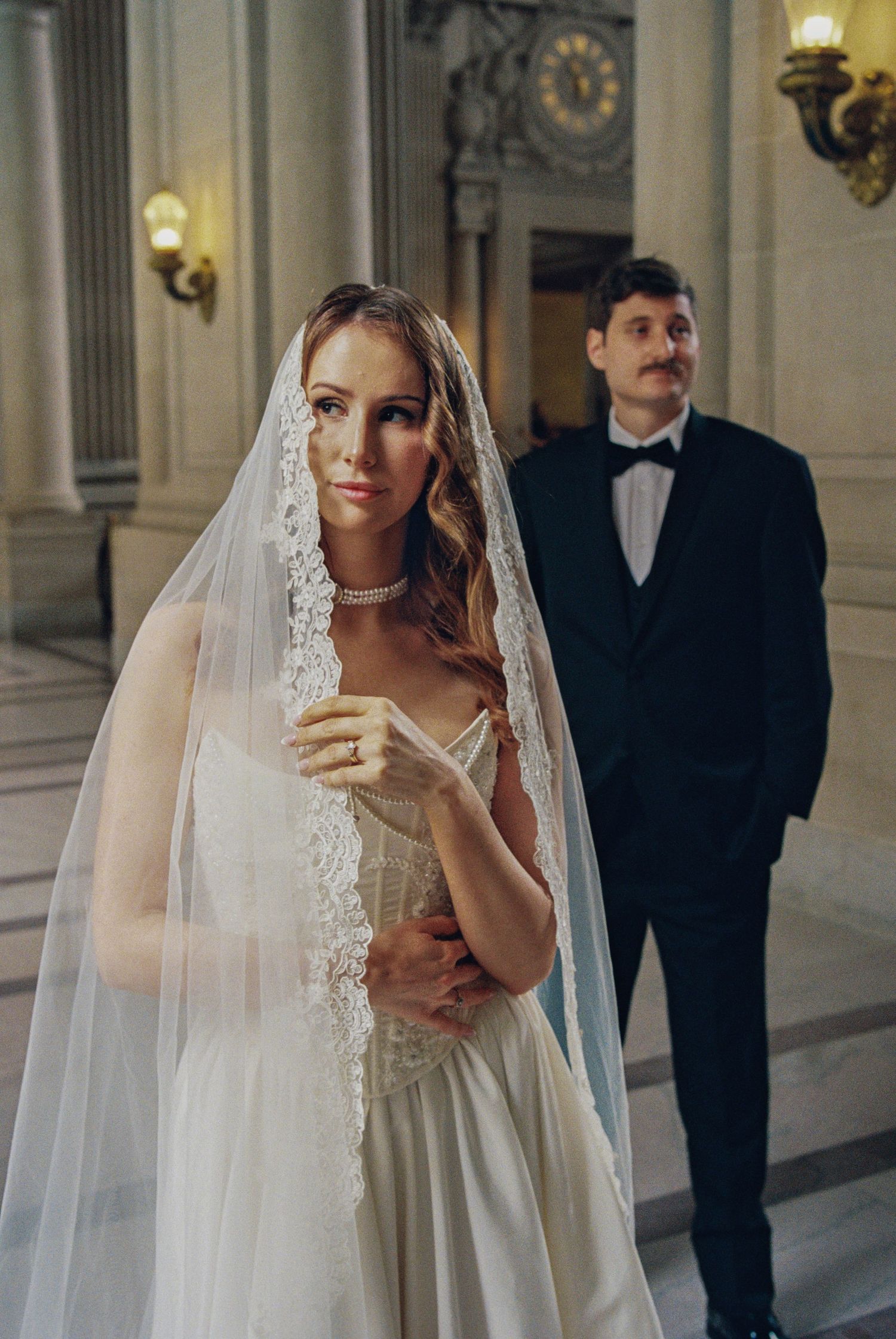 Bride in lace veil and white gown stands elegantly with groom in black tuxedo at a grand wedding venue.