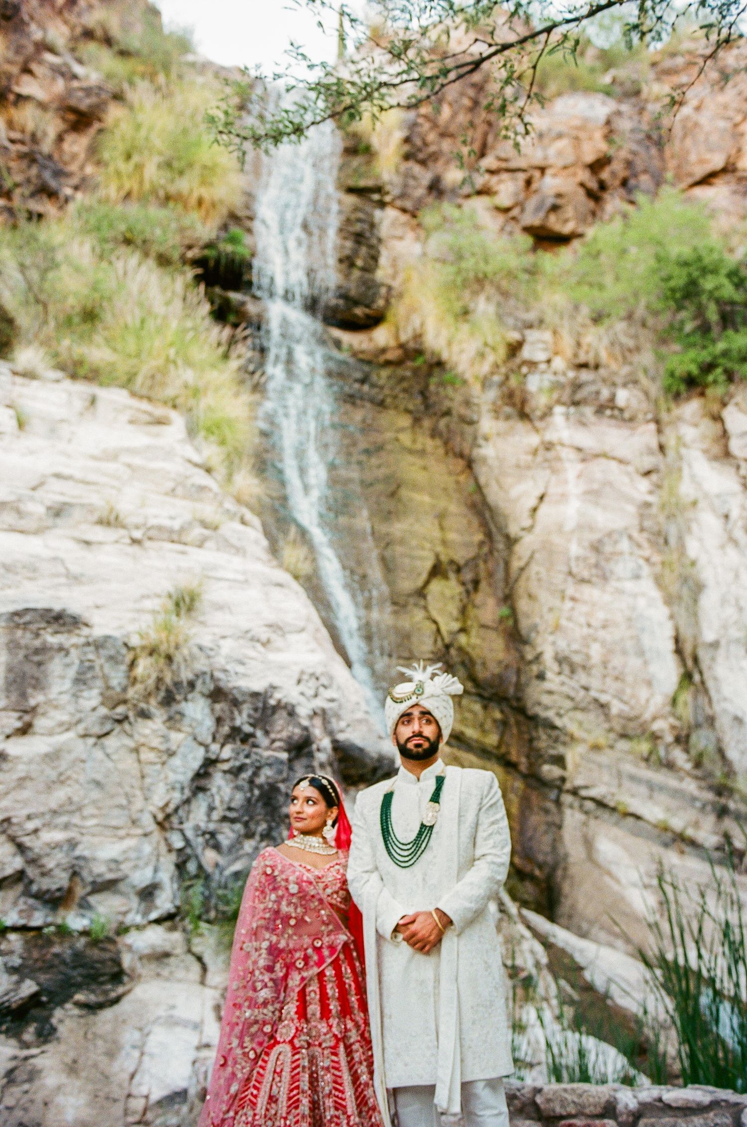 A couple in traditional wedding attire poses against a scenic waterfall backdrop.