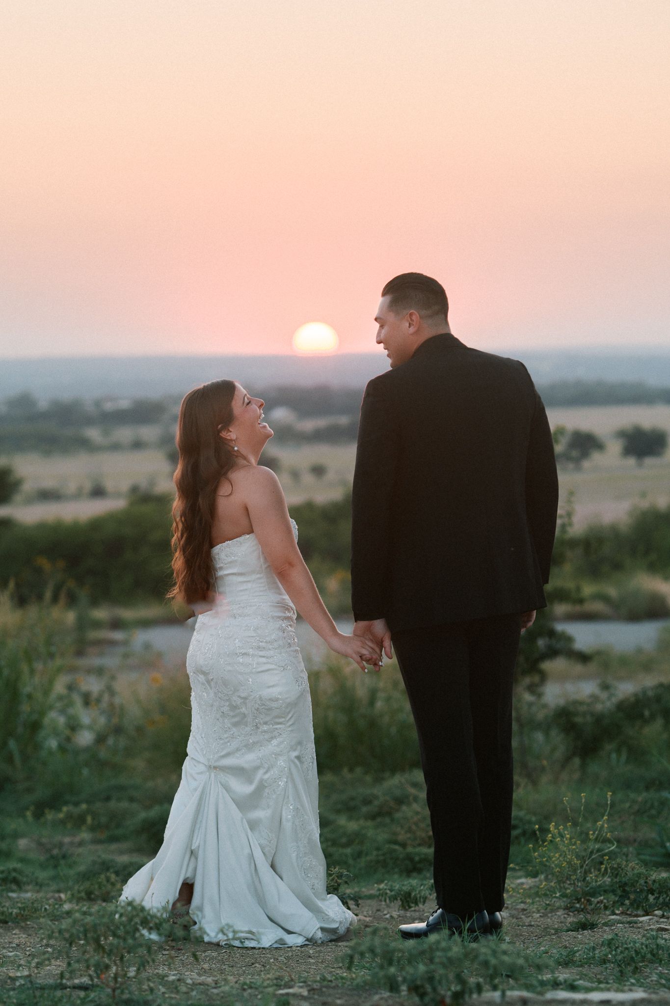 Bride and groom hold hands gazing at each other against a stunning golden sunset over open countryside.