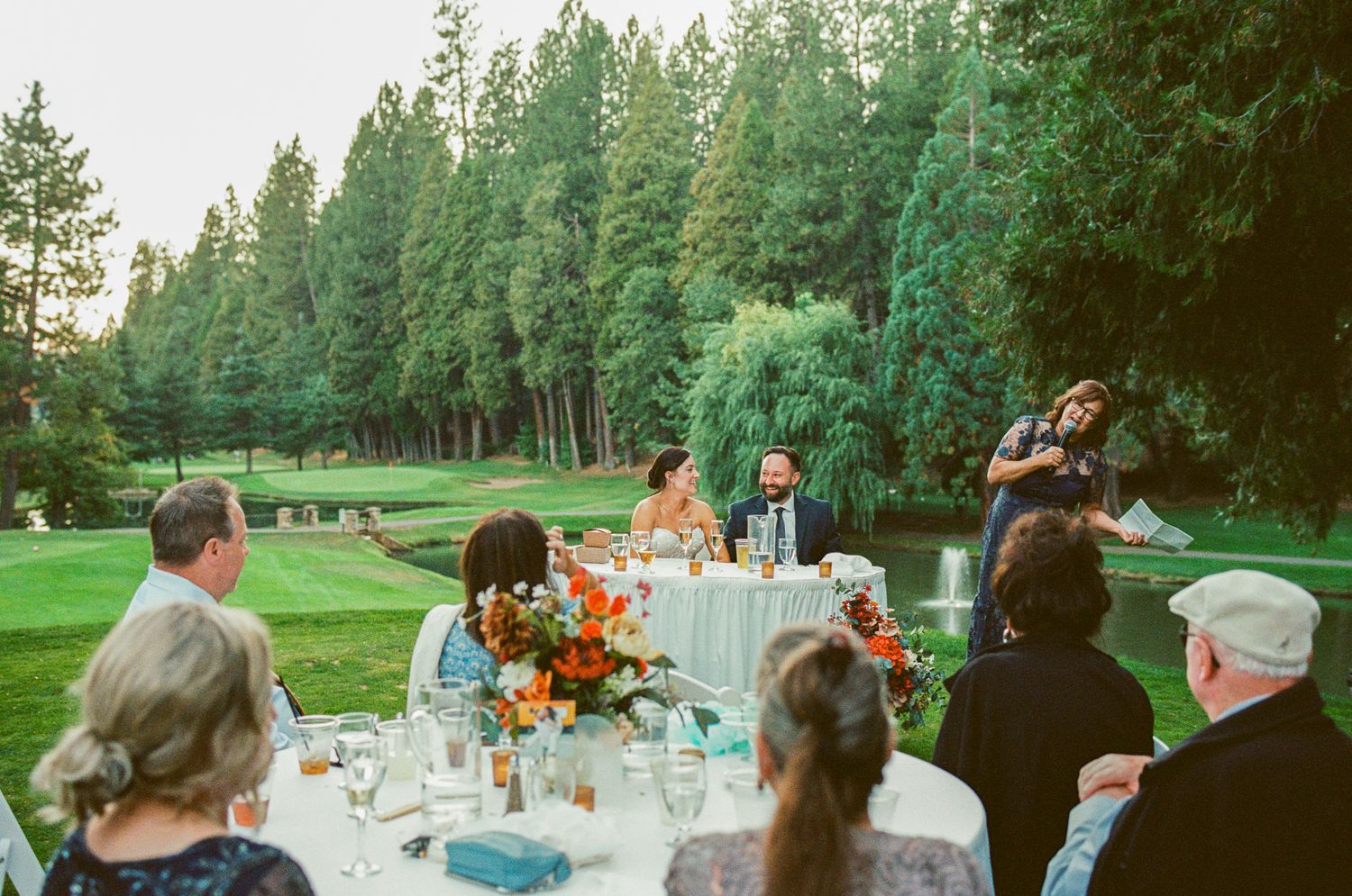 Outdoor wedding reception on a golf course with guests seated at a floral table while someone gives a toast.