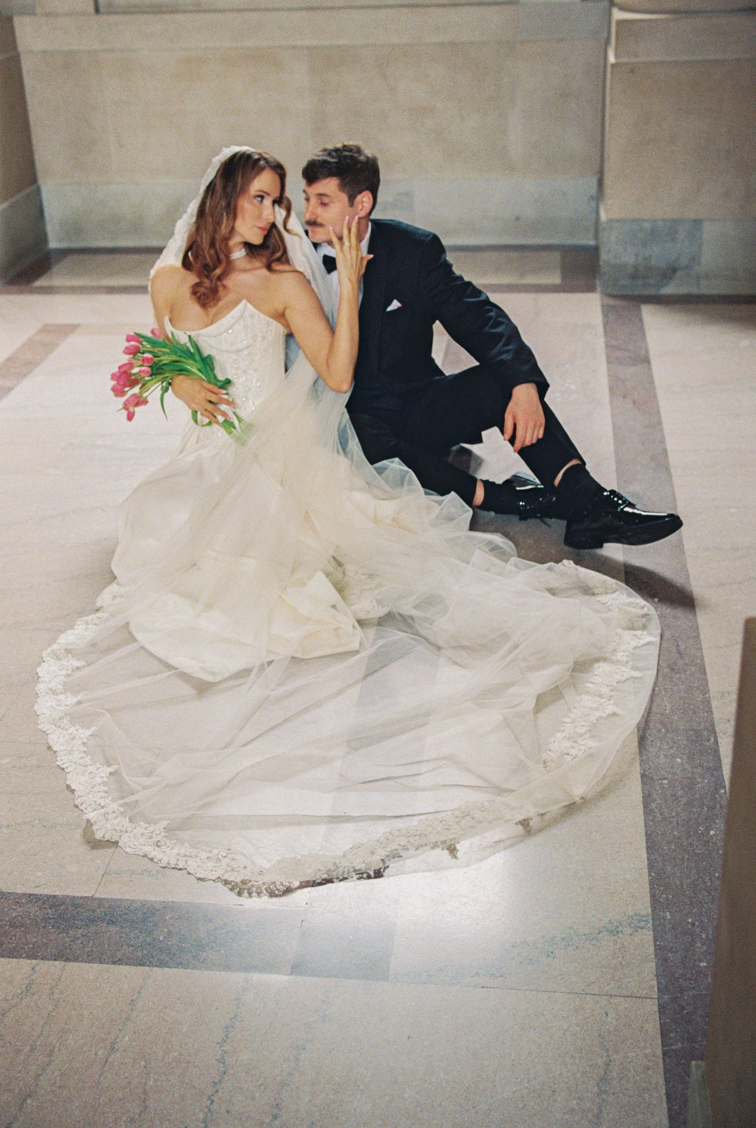 Bride in white gown with lace train holds tulips beside groom in black tuxedo sitting on marble floor.