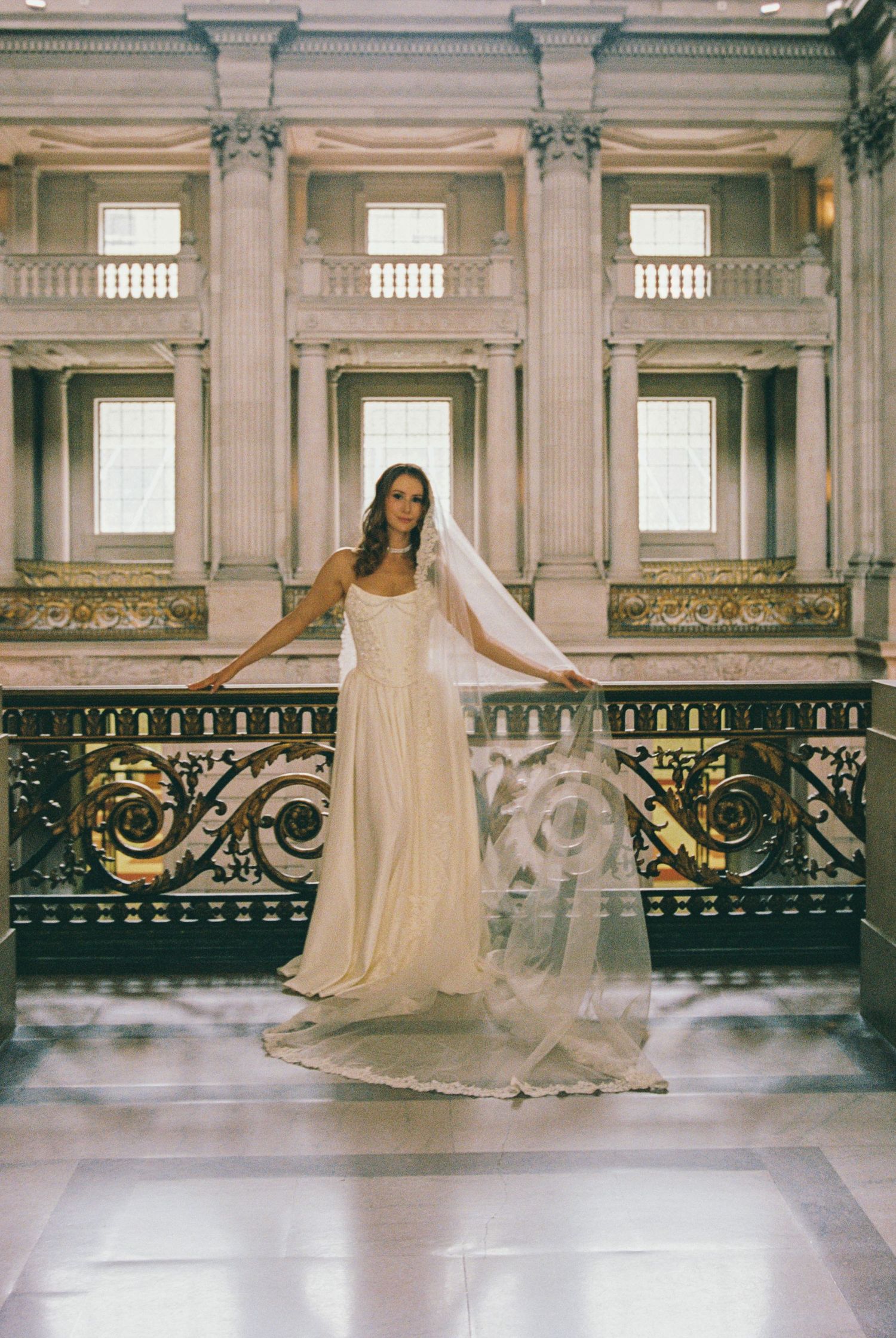 Bride in elegant white gown and veil poses on ornate balcony inside grand neoclassical building.