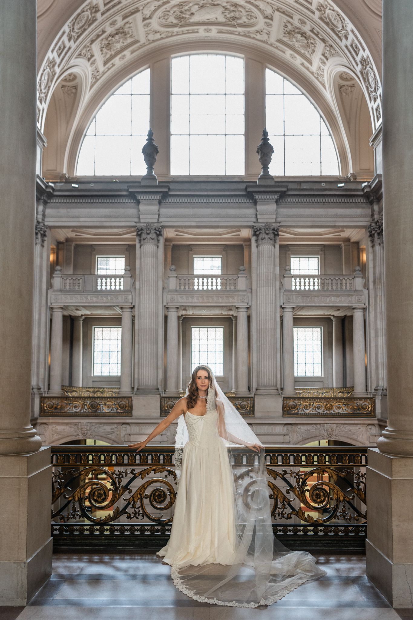 Bride in white gown and veil poses on ornate balcony inside San Francisco City Hall grand rotunda.