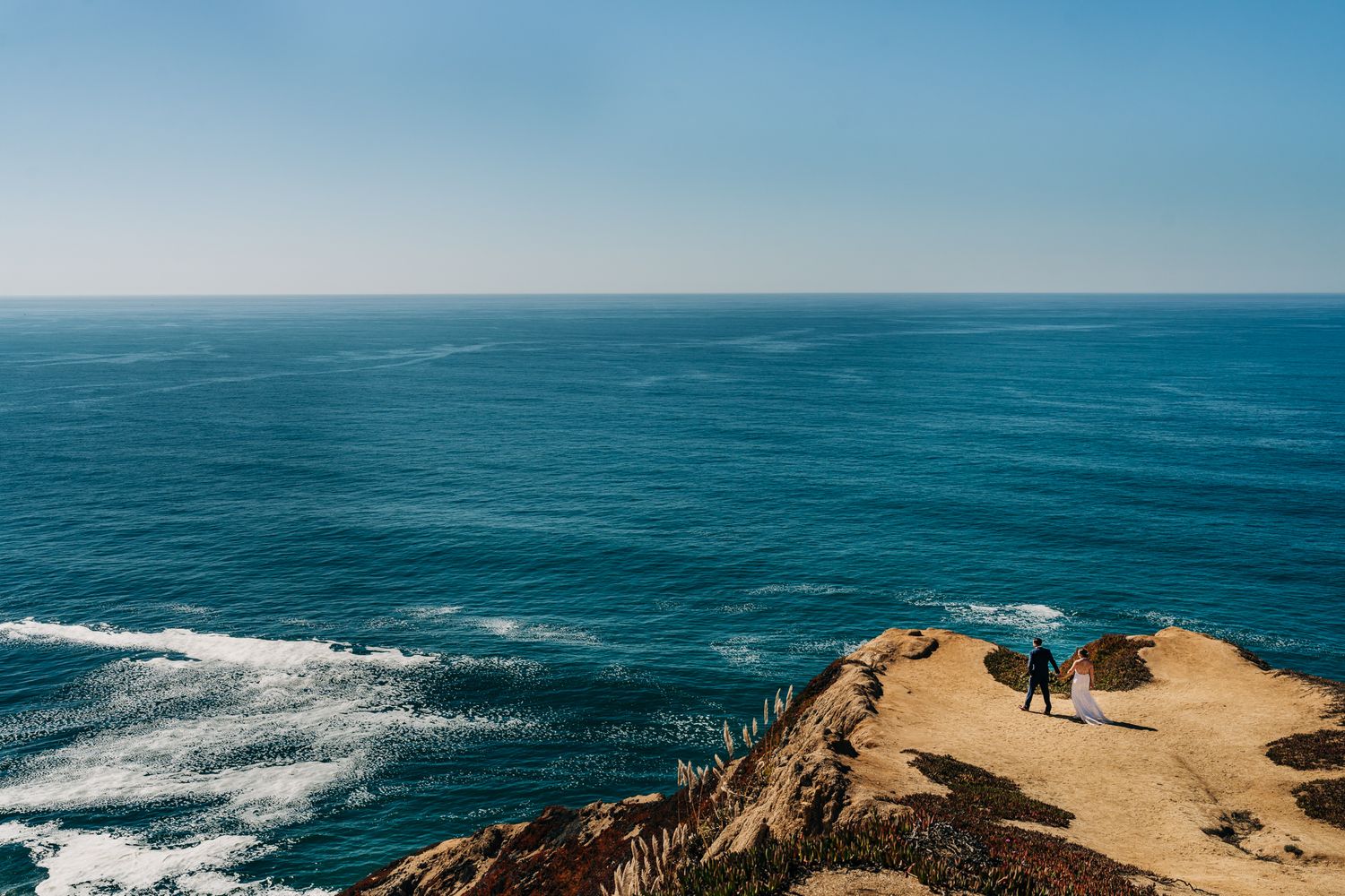 Aerial view of people standing on a coastal cliff overlooking the vast blue Pacific Ocean on a sunny day.