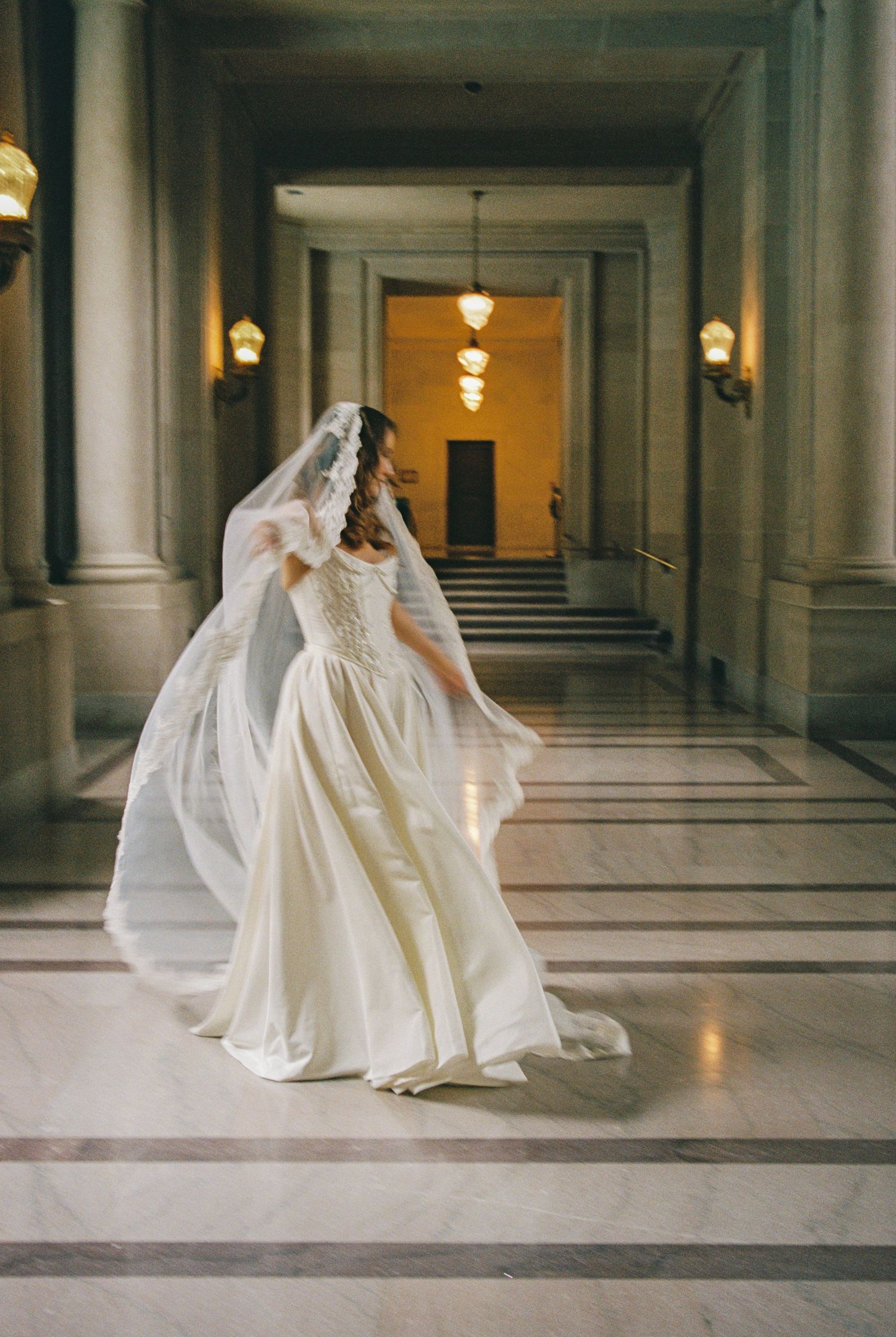 Bride in flowing white gown and veil twirls in elegant marble hallway with warm chandelier lighting.