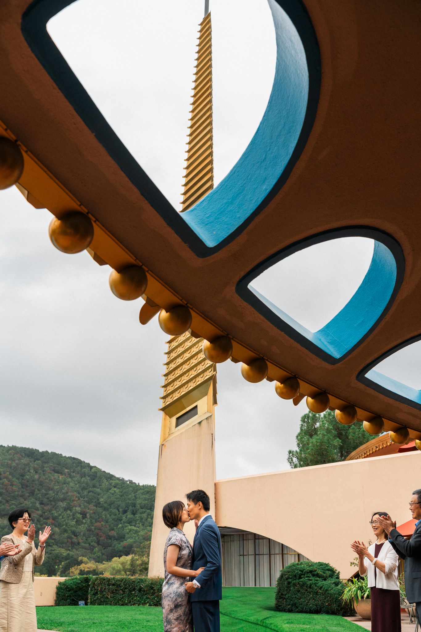 Couple shares first kiss under ornate curved architecture with golden spire, guests applauding, green hills backdrop.
