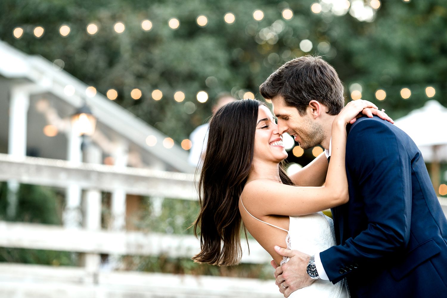Romantic wedding couple embracing under twinkling string lights at dusk.