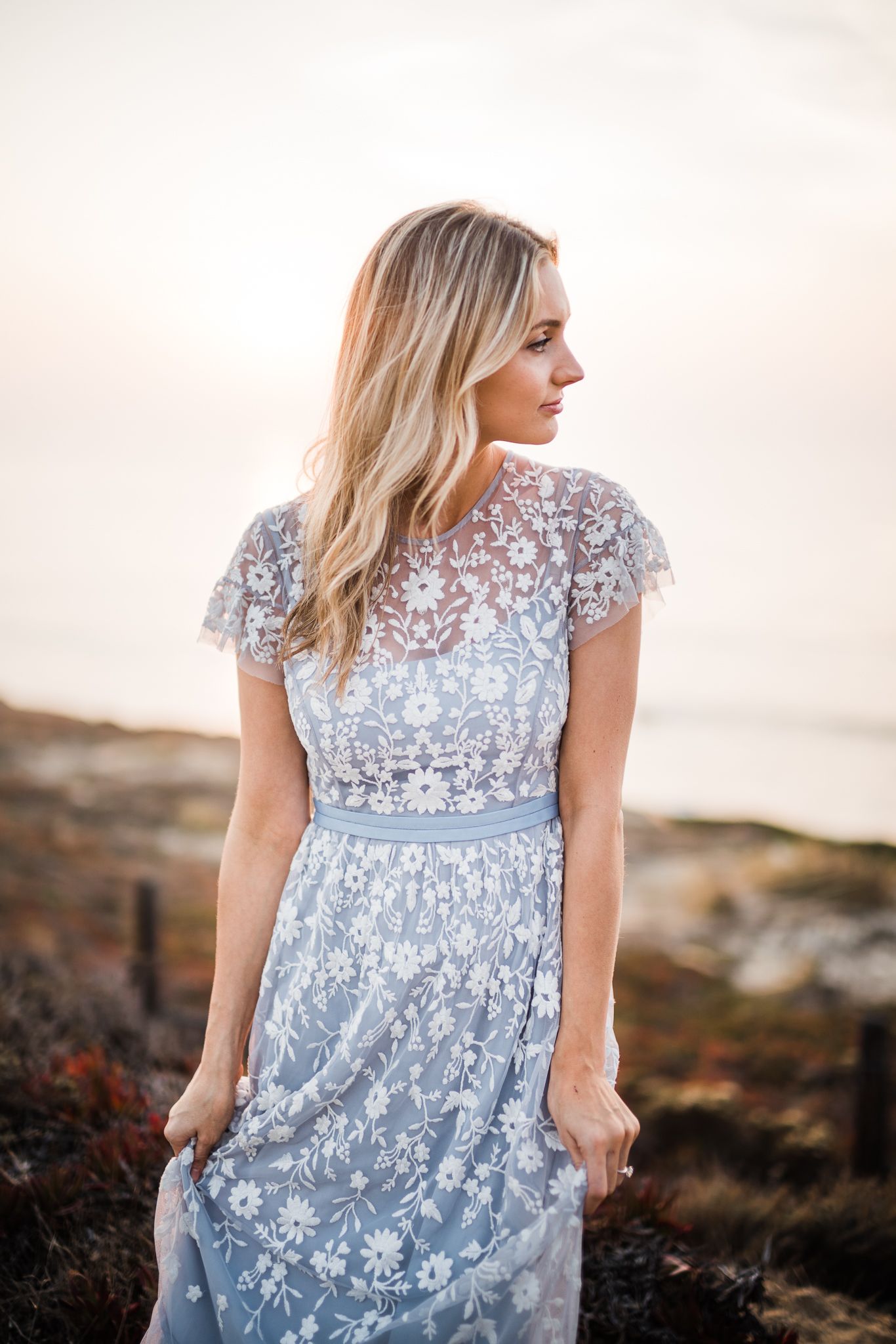 Woman in light blue lace dress poses against sunset backdrop in romantic portrait series.