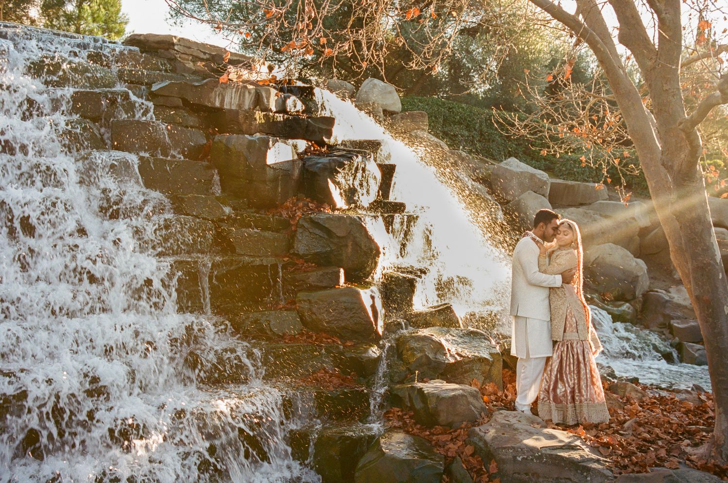 Couple sharing a romantic kiss beside a cascading stone waterfall surrounded by autumn foliage in golden sunlight.