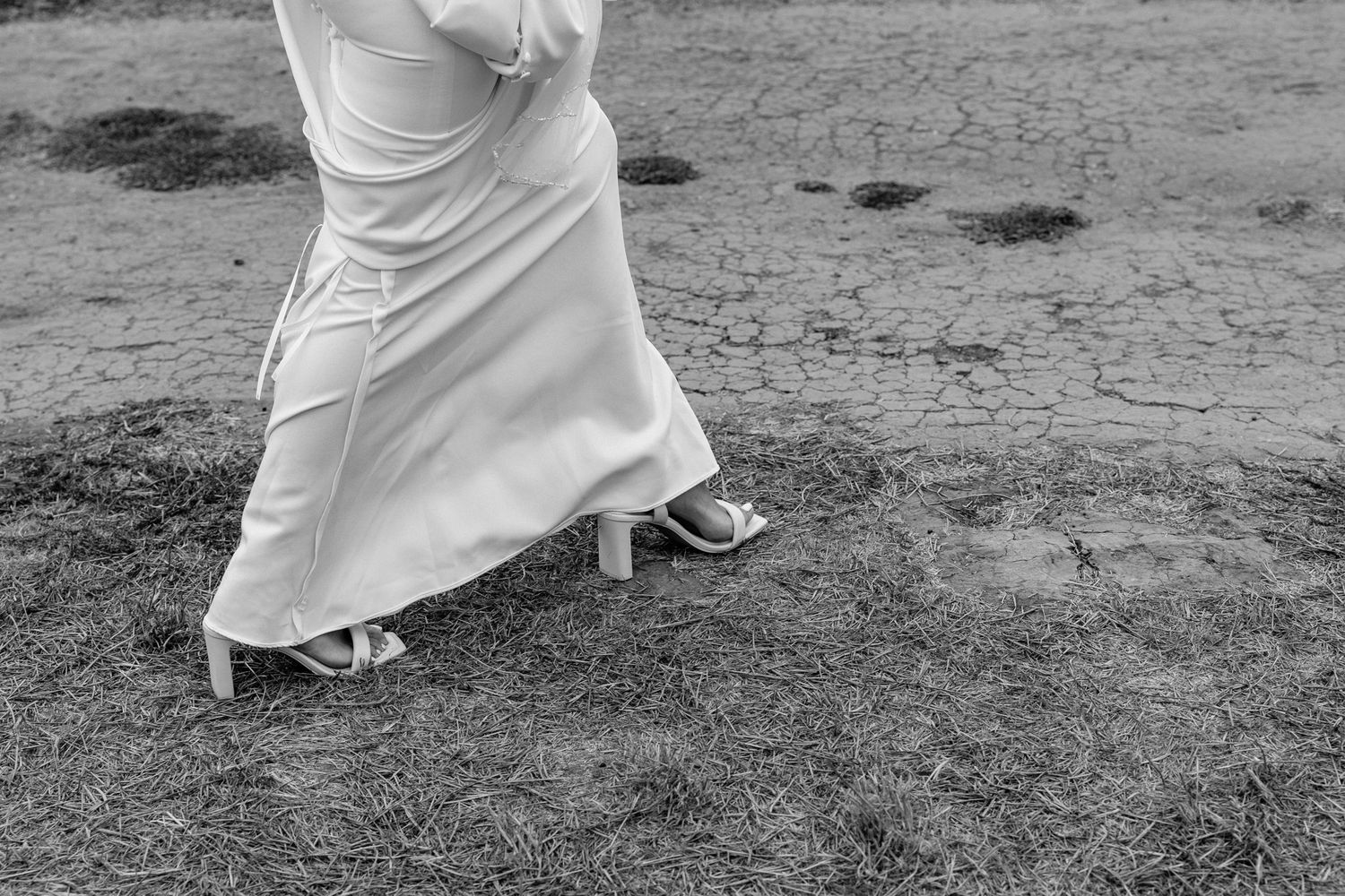 Close-up of flowing white wedding dress and shoes on grassy terrain.