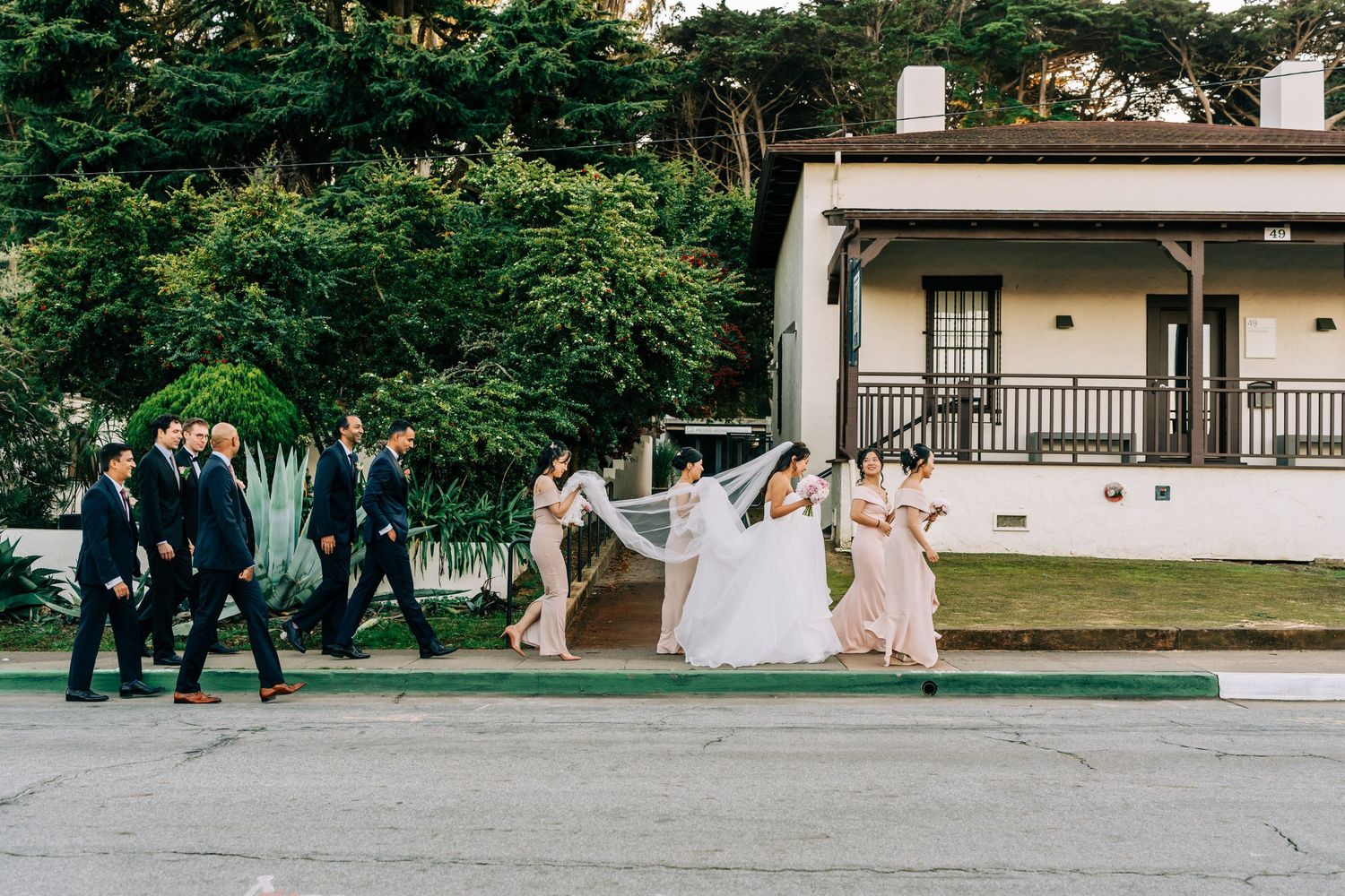 Wedding party members walk in a line across a green lawn in front of a white building.
