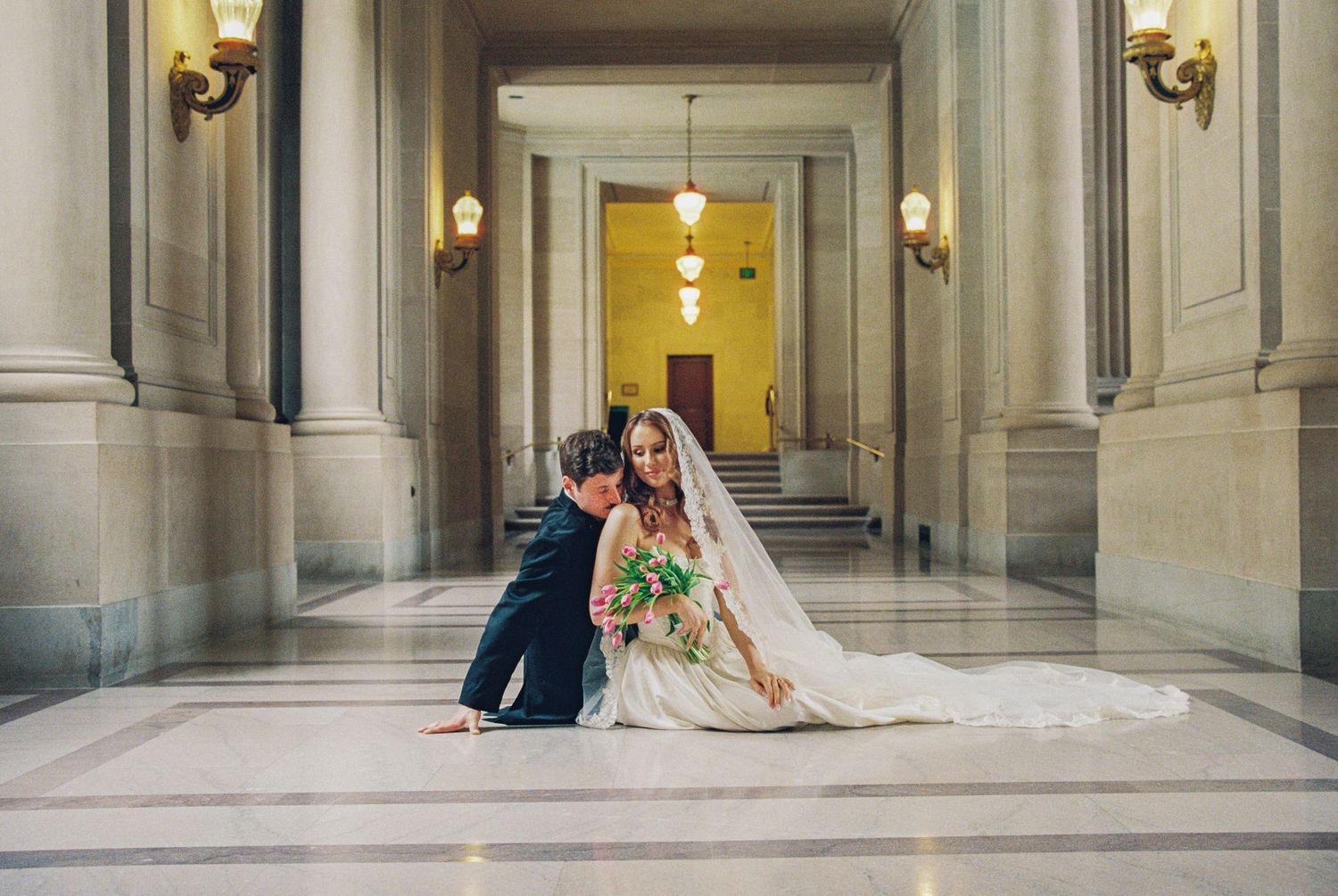 Bride and groom sitting on marble floor in elegant hall, bride in white gown with veil and bouquet.