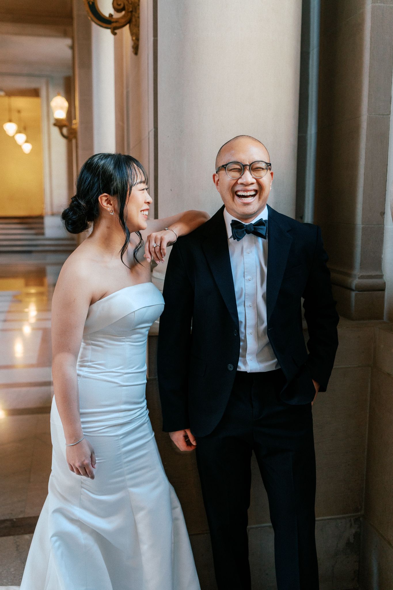 Bride in strapless white gown and groom in black tuxedo laughing together against marble columns.