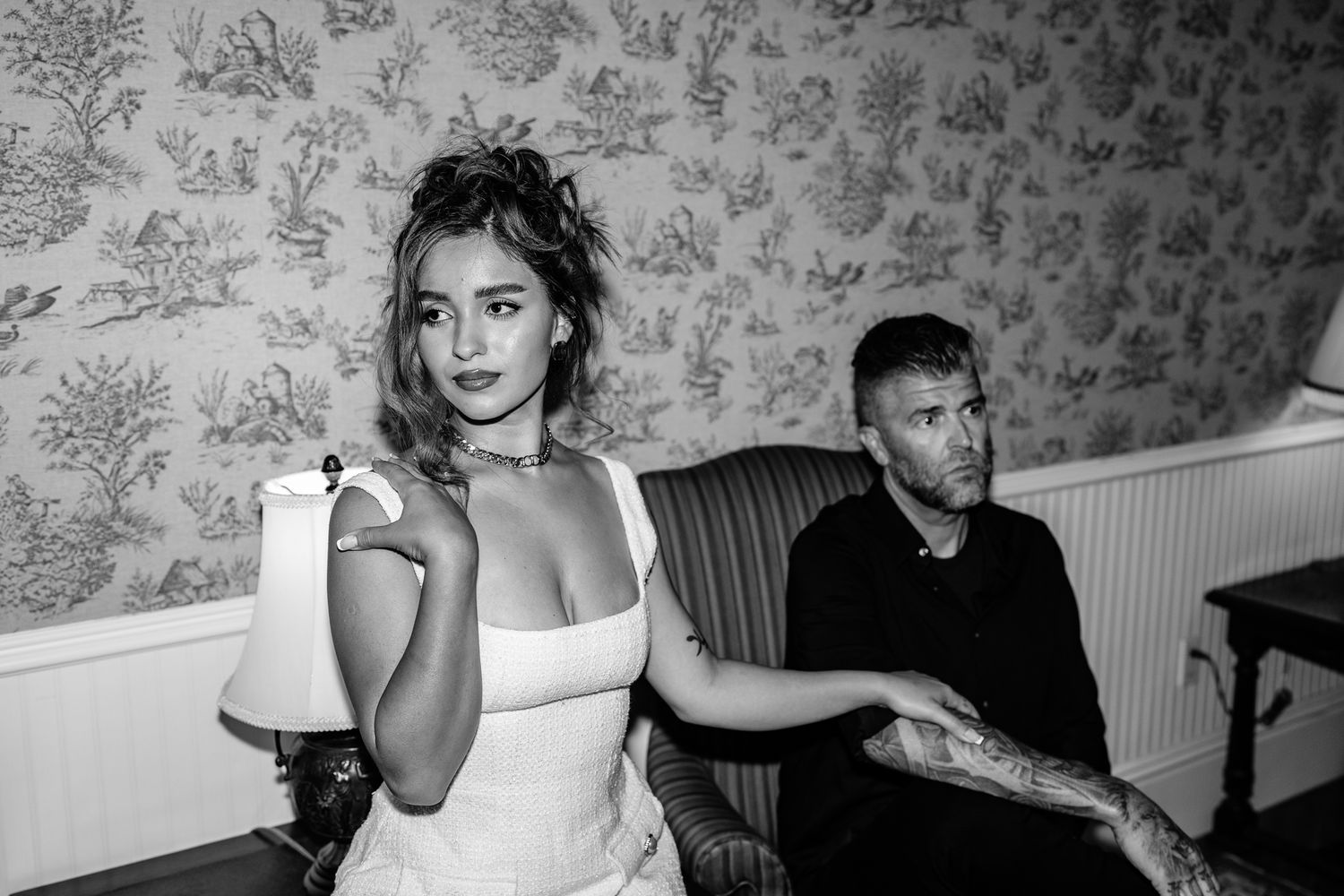 Black and white photo showing two people in a room with floral wallpaper, one in a white dress and one seated on a couch.