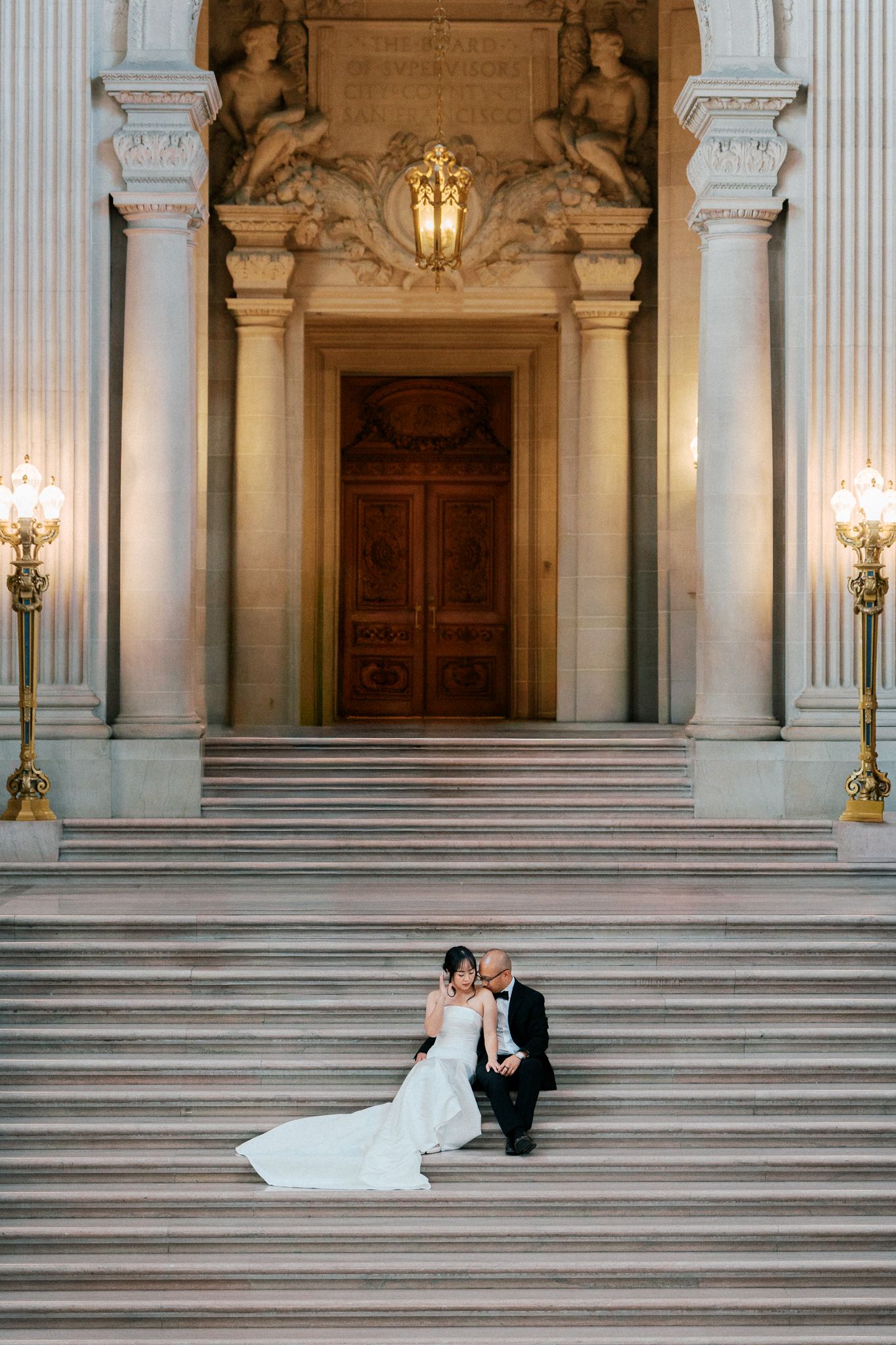 Bride and groom sit together on grand marble steps inside San Francisco City Hall, surrounded by ornate columns and warm lighting.