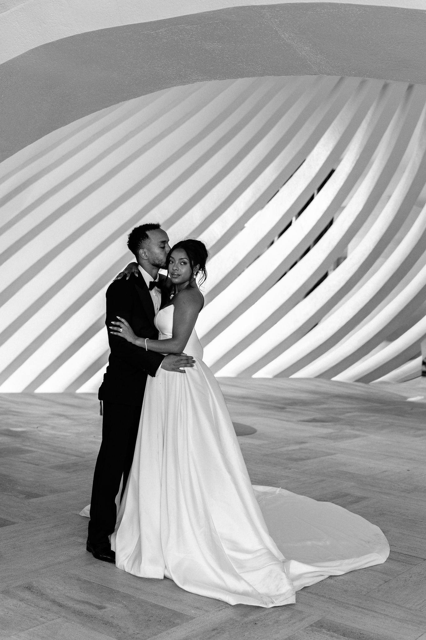 Black and white photo of a couple sharing a romantic embrace in wedding attire against a modern architectural backdrop.