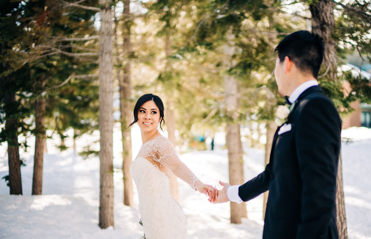 A romantic winter wedding scene with a bride and groom holding hands among snow-covered pine trees.