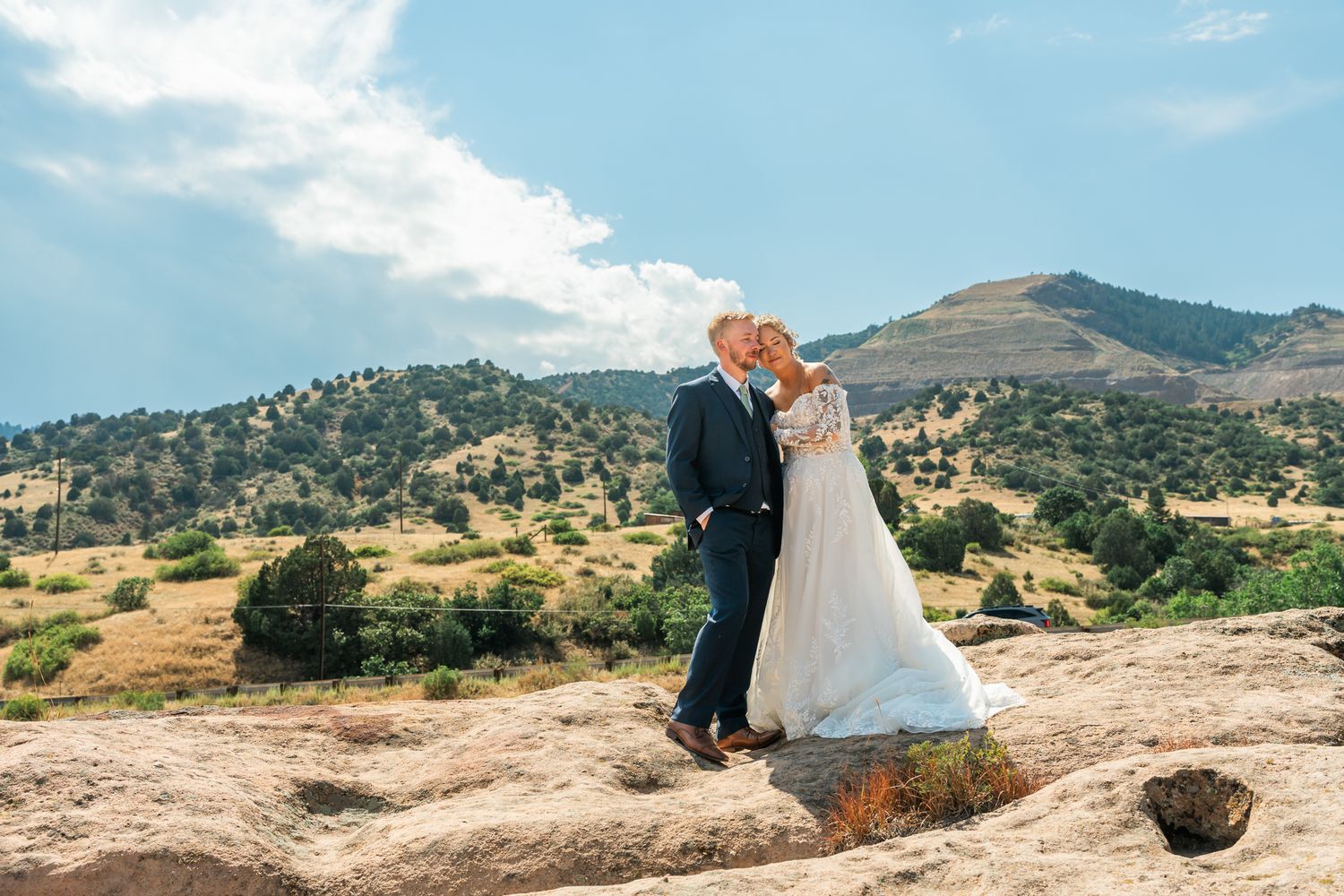 Bride and groom share a romantic kiss on rocky terrain with scenic mountain landscape and blue sky backdrop.