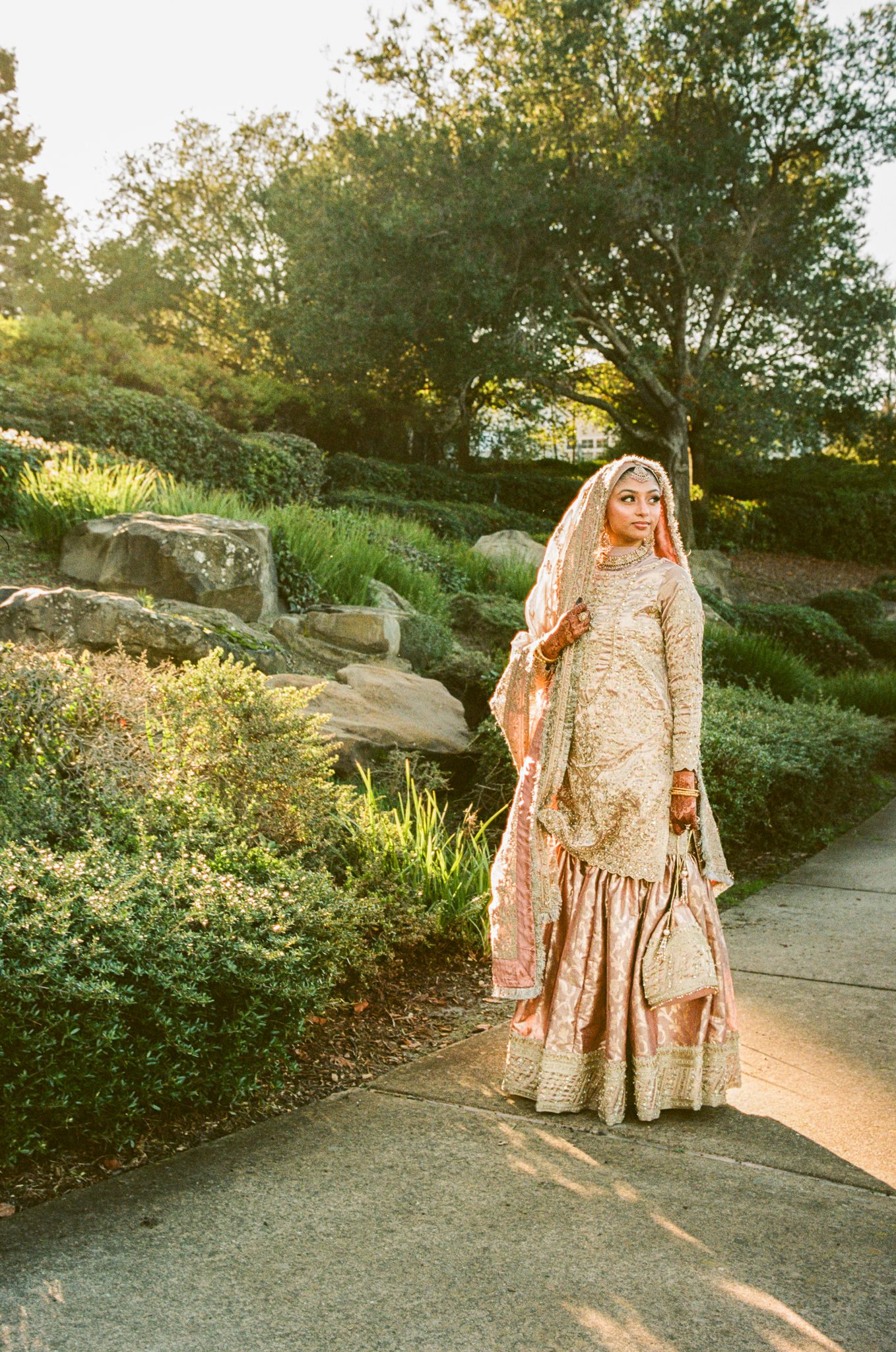 Traditional South Asian bride in peach lehenga poses outdoors during golden hour sunset photoshoot.