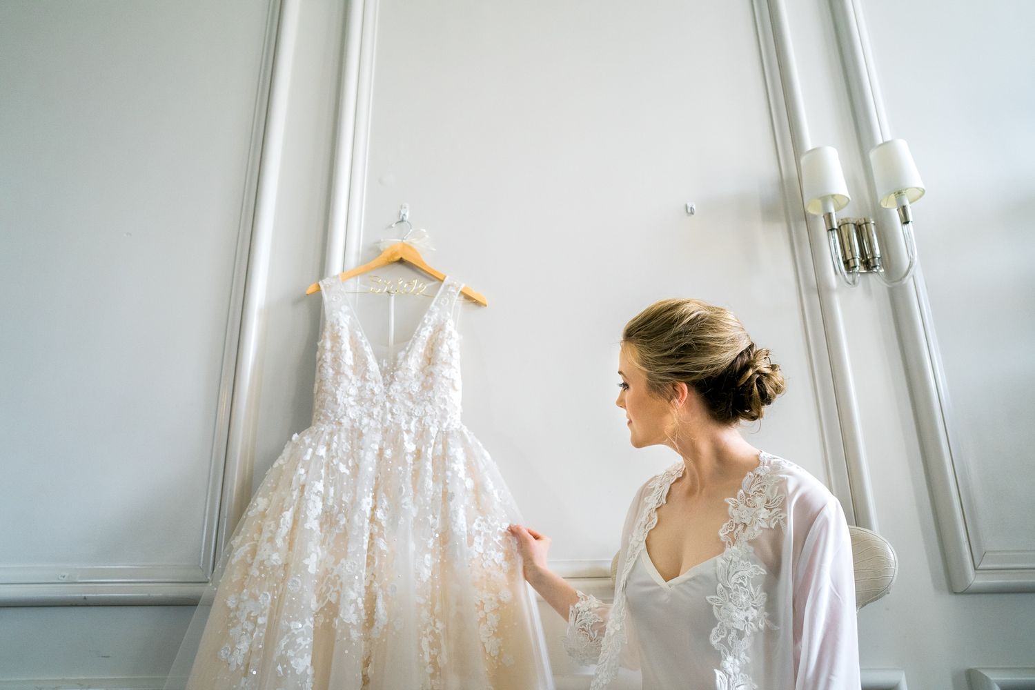 Elegant white lace wedding dress hanging in bright room with minimalist decor.