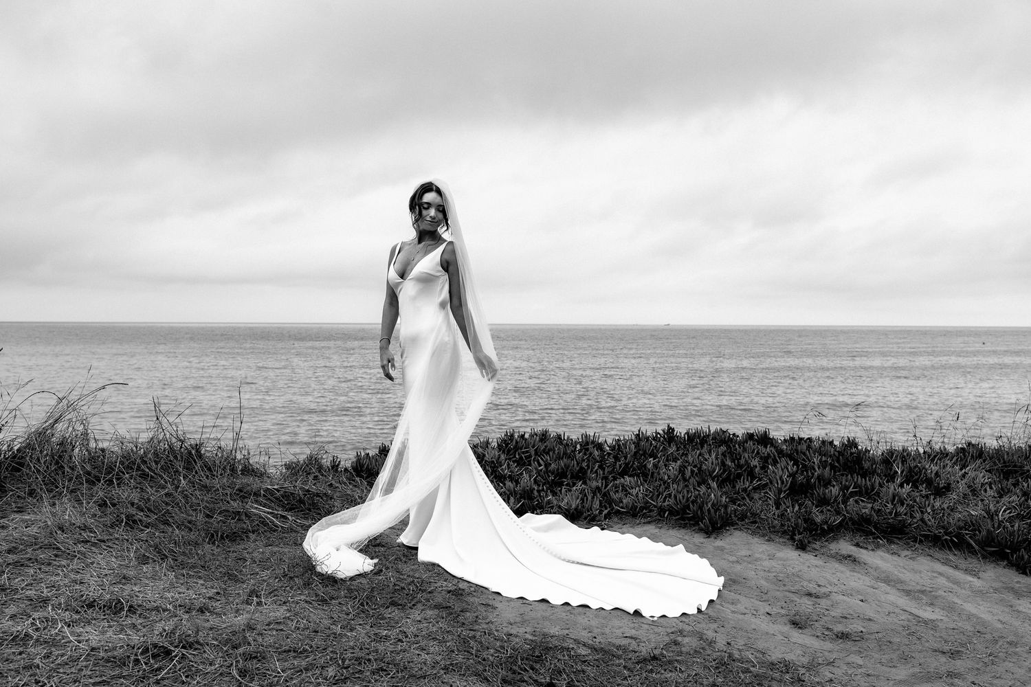 Elegant bridal portrait with flowing train against dramatic coastal scenery.