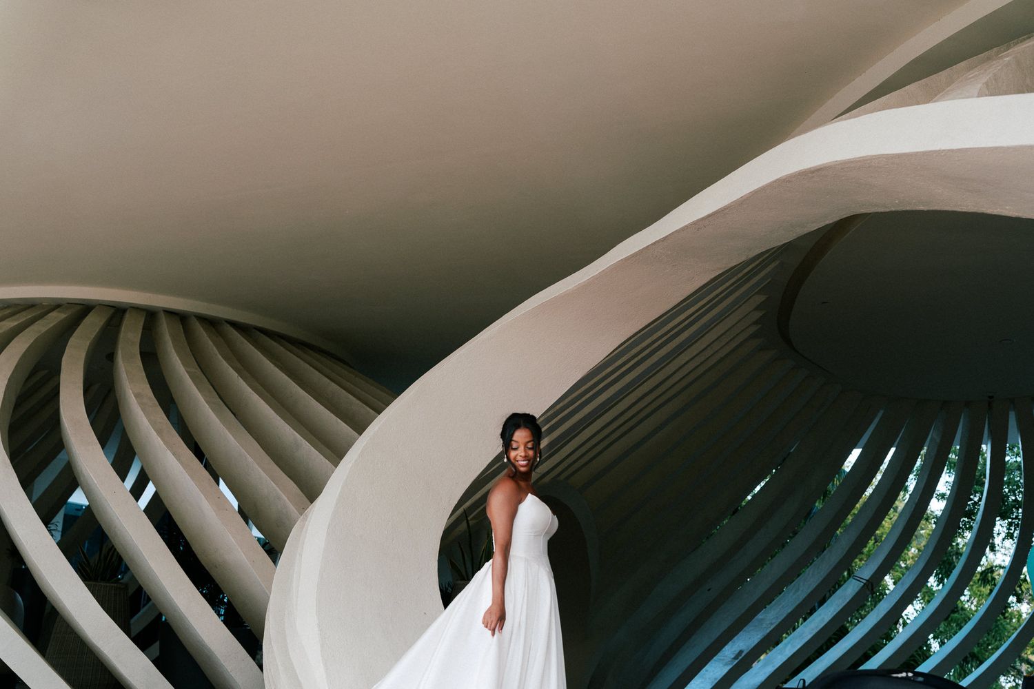 Bride in white gown poses on spiral staircase of modern architectural building with curved concrete walls.