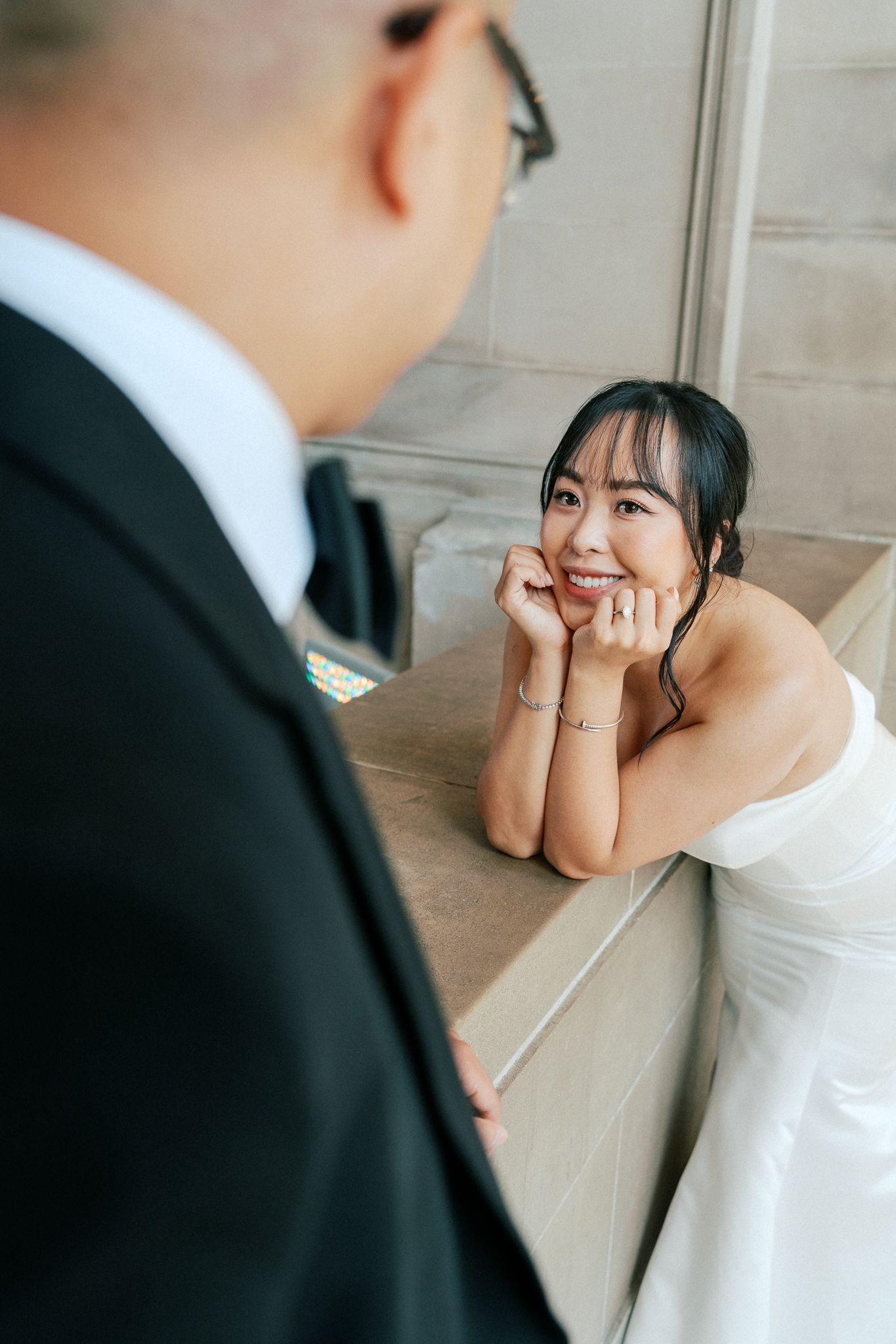 Smiling Asian bride in white strapless gown rests chin on hands, gazing up at groom in black suit.