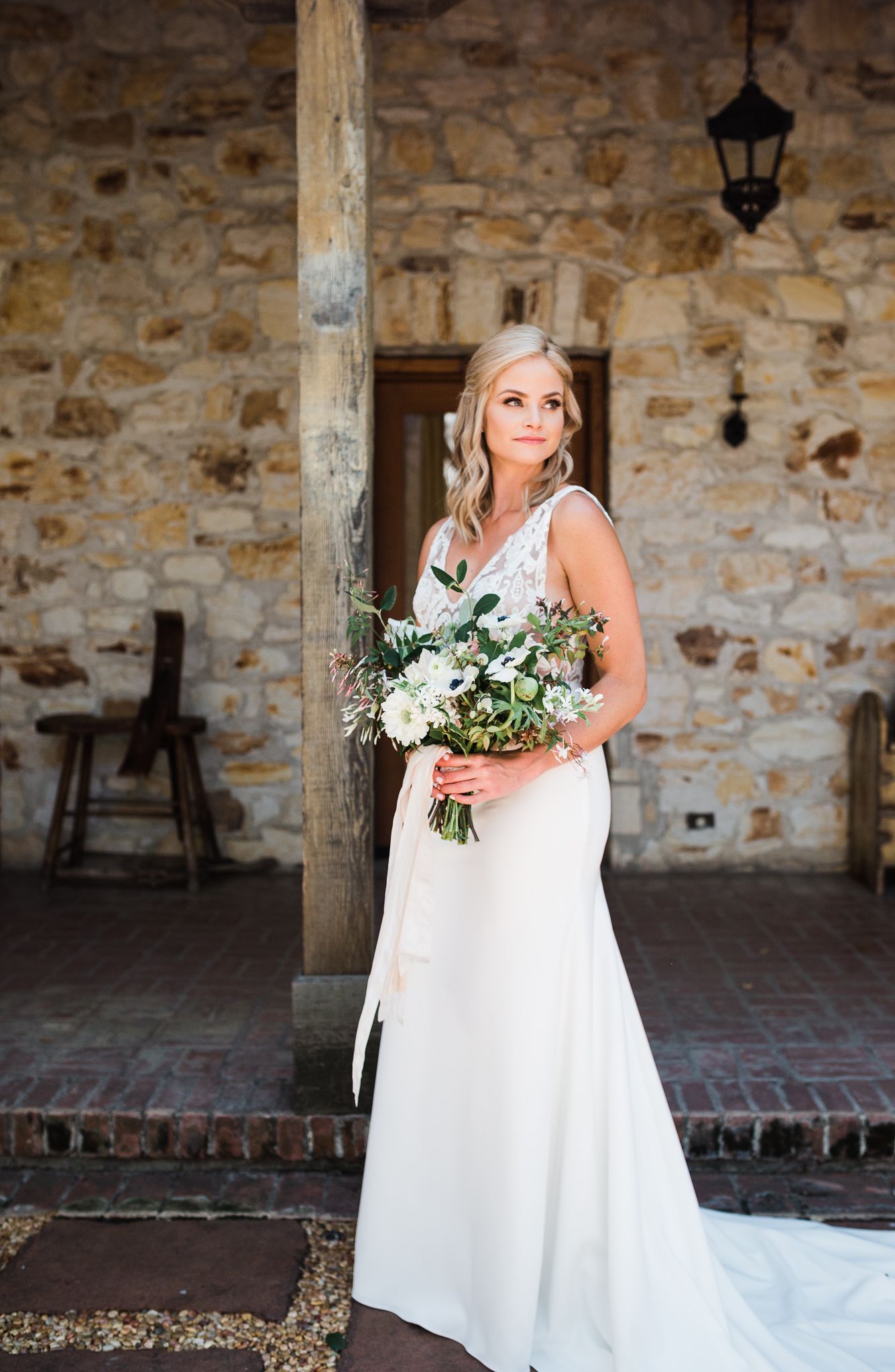 Bride in elegant white dress holding a fresh white and green bouquet against stone wall backdrop.