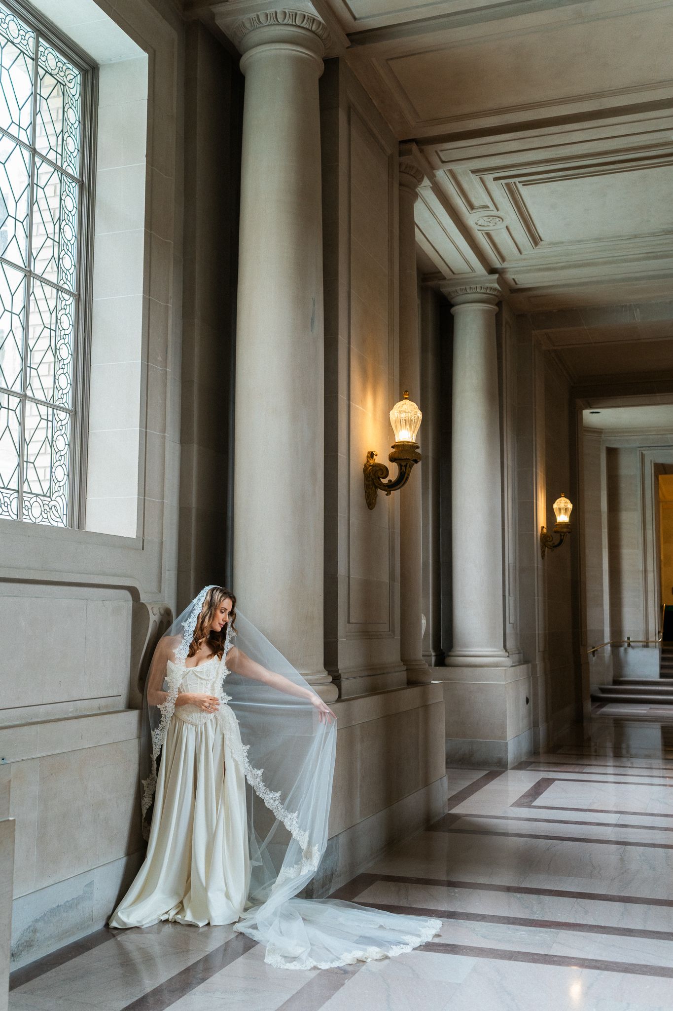 Elegant bride in flowing white gown and cathedral veil stands among marble columns in SF City Hall.