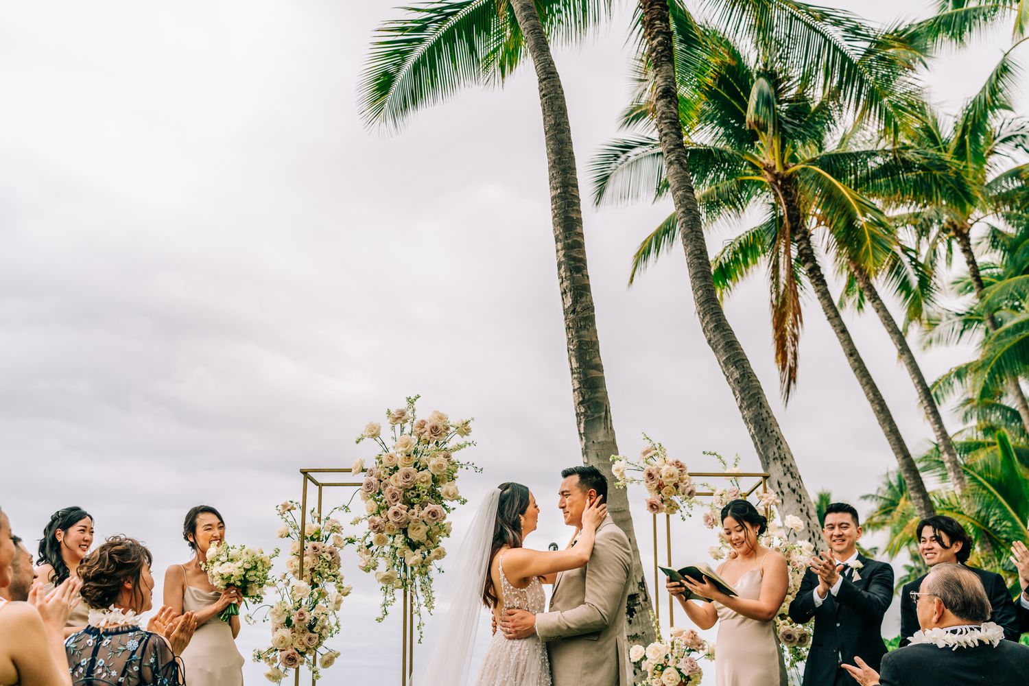 Outdoor tropical wedding ceremony with couple exchanging vows under floral arch, palm trees, and overcast sky.