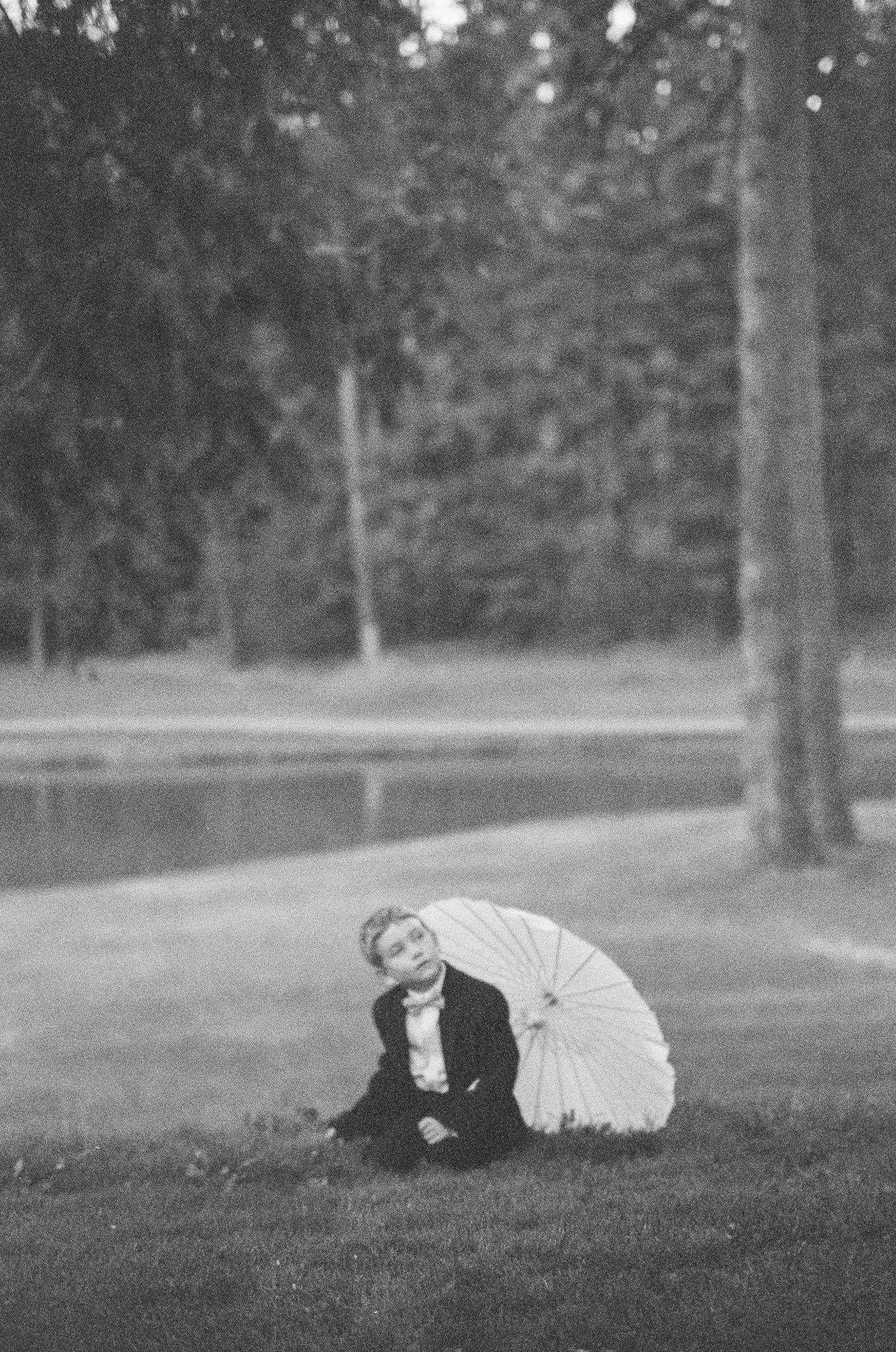 Black and white vintage photo of a child sitting on grass holding a parasol umbrella near a pond and trees.