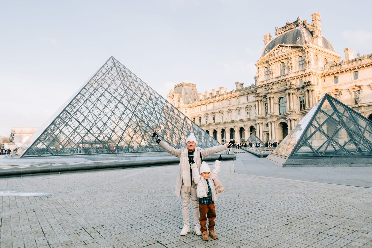 Tourists pose in front of the iconic glass pyramid at the Louvre Museum in Paris during golden hour.