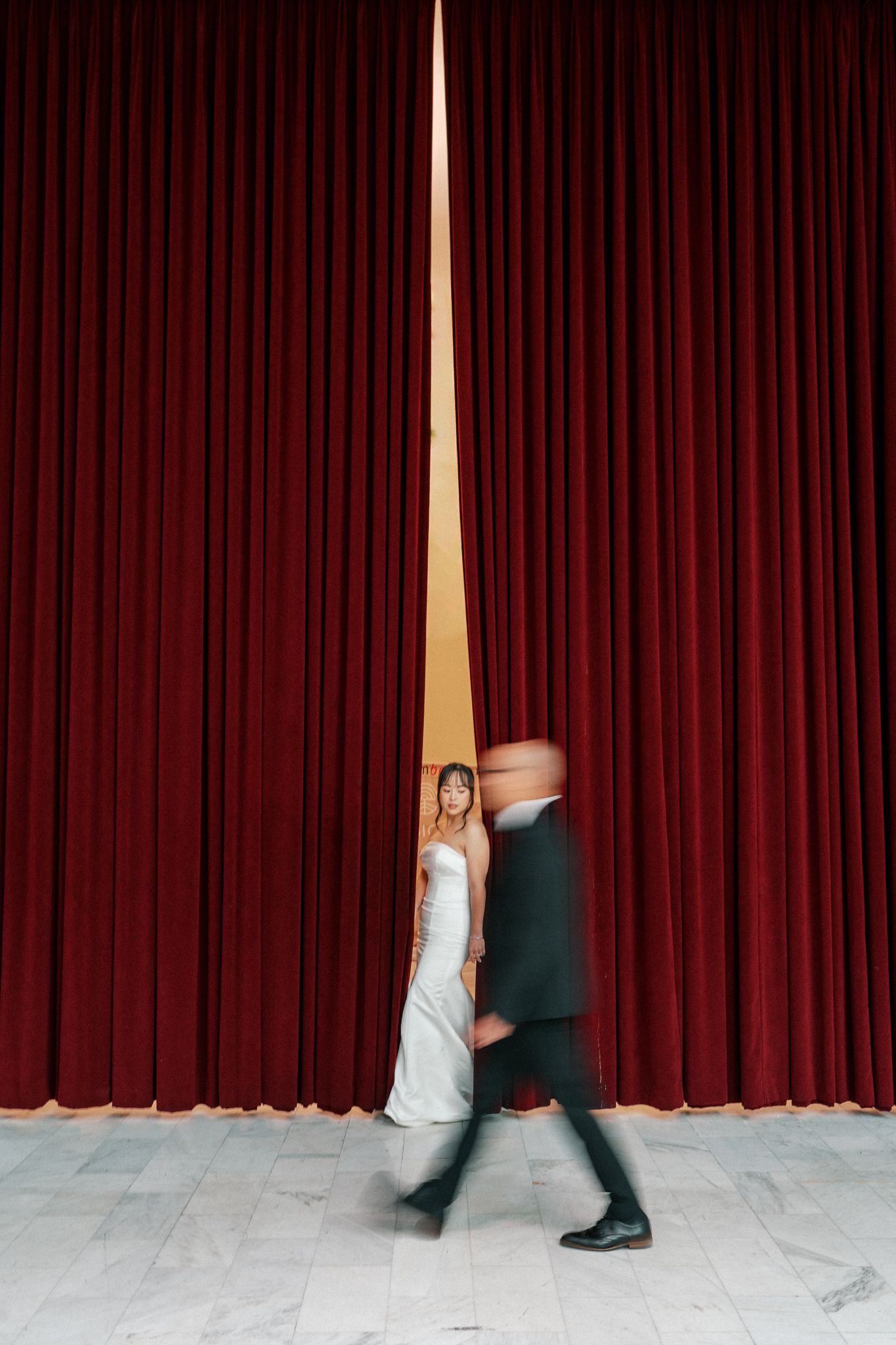 Woman in white gown stands between red velvet curtains as a blurred figure walks past on marble floor.