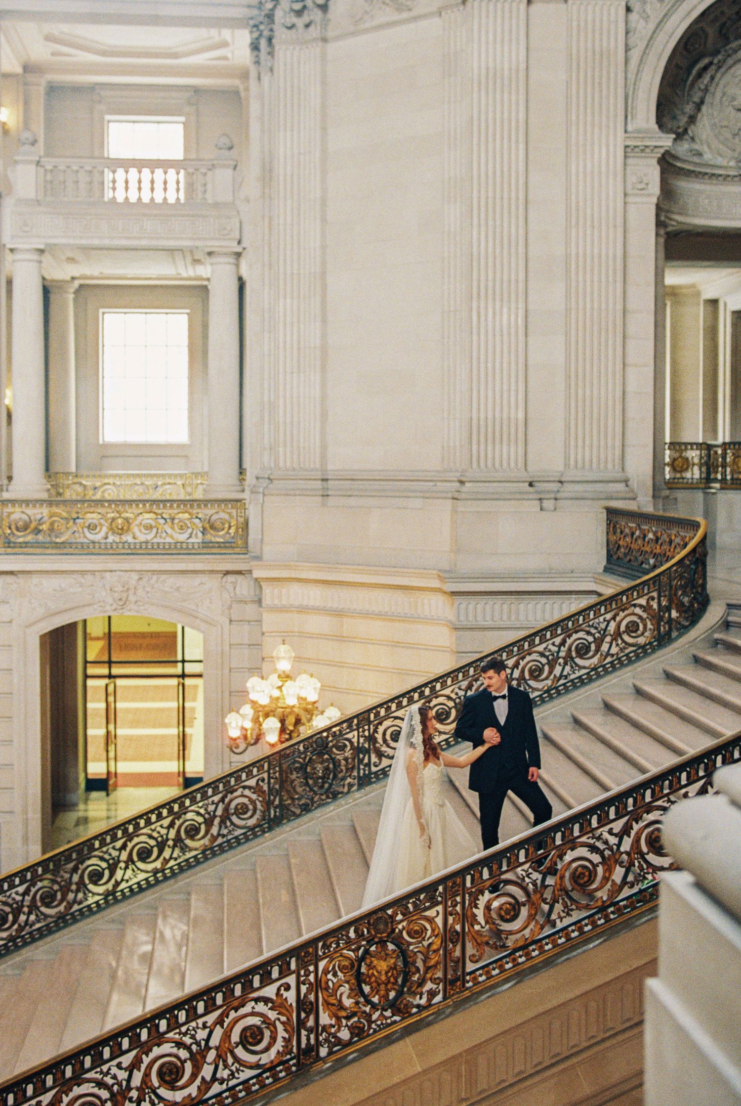 Bride and groom walk down ornate grand staircase at San Francisco City Hall during elegant wedding ceremony.