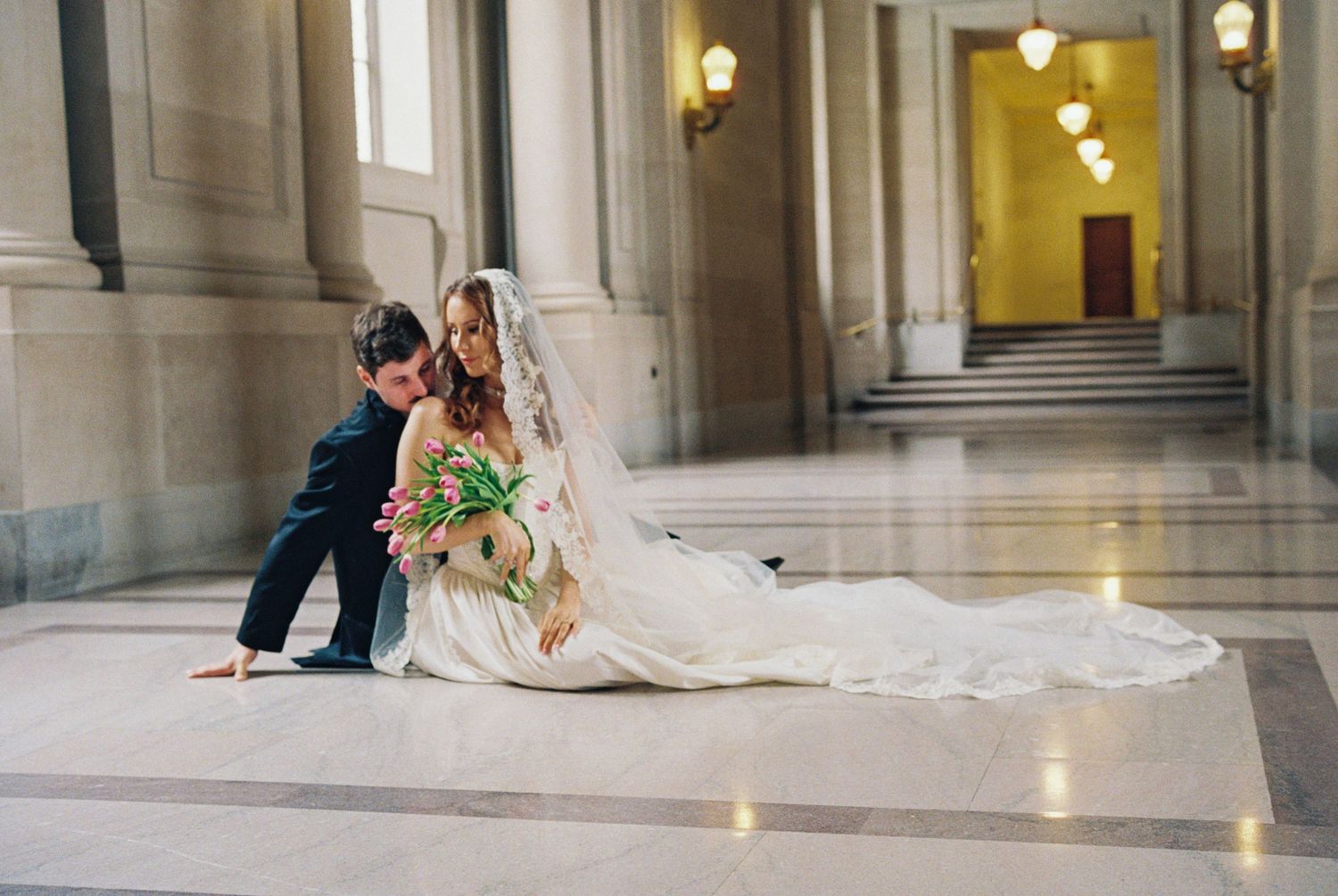 Bride and groom sitting on marble floor in elegant hall, bride in white gown with veil holding pink tulips.