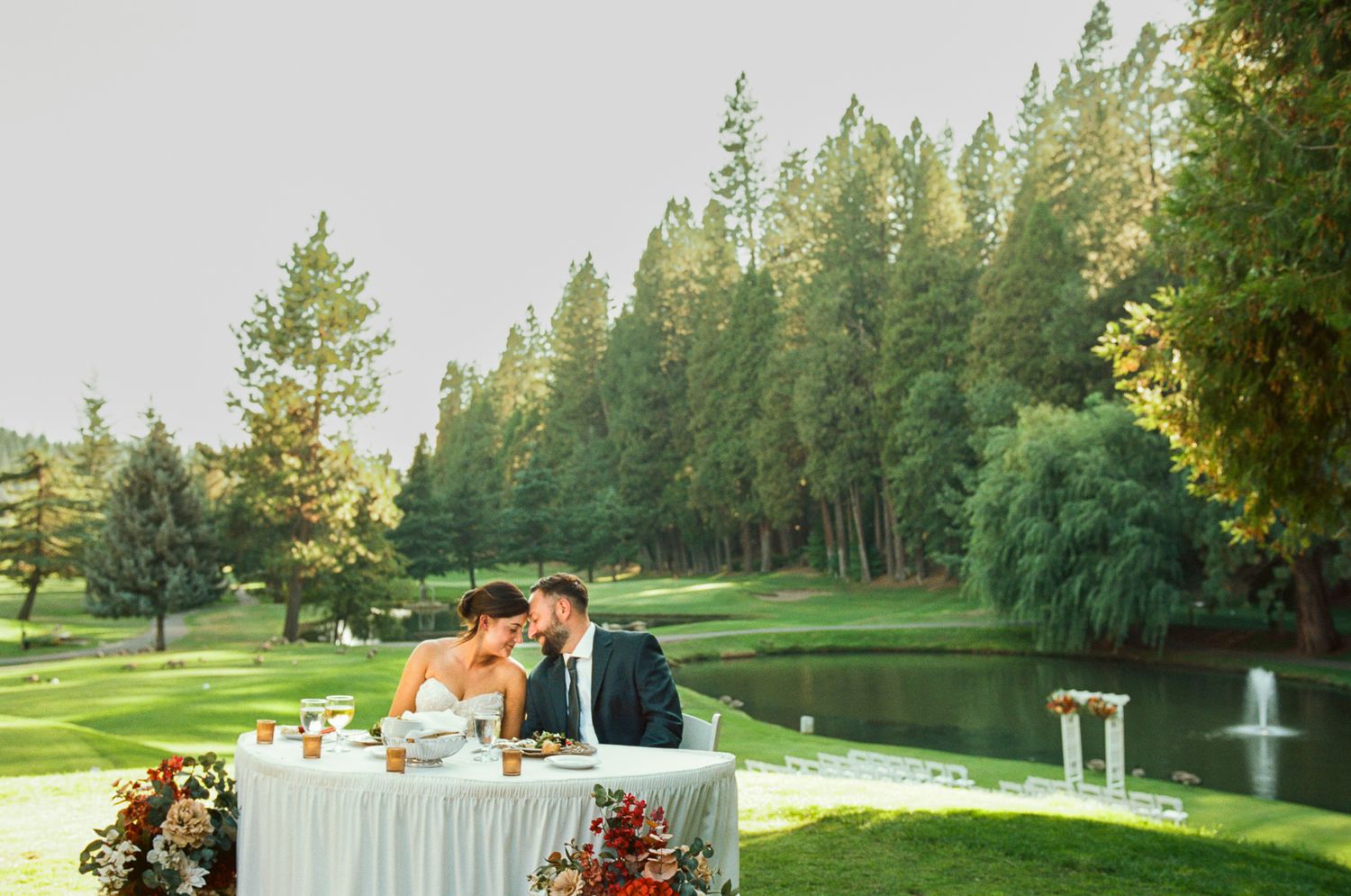 Wedding ceremony setup on a lush golf course with elegant table decor and pine trees in the background.