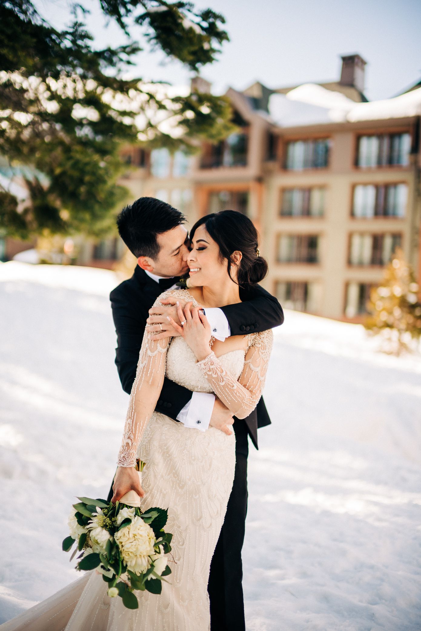 A romantic winter wedding photo with a couple embracing in the snow outside a mountain resort.