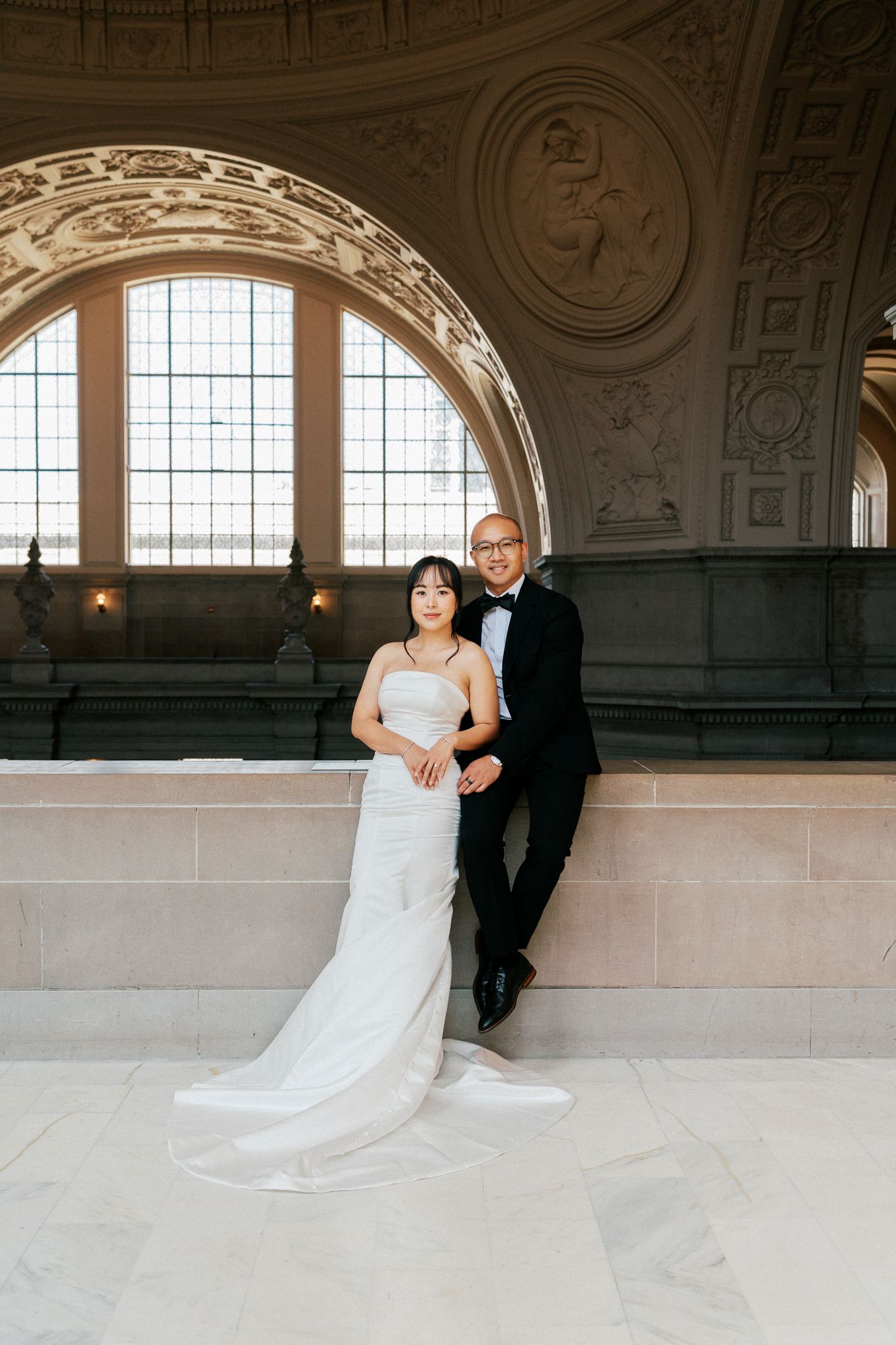 A bride in a white gown and groom in a tuxedo pose elegantly inside a grand ornate hall with arched windows.