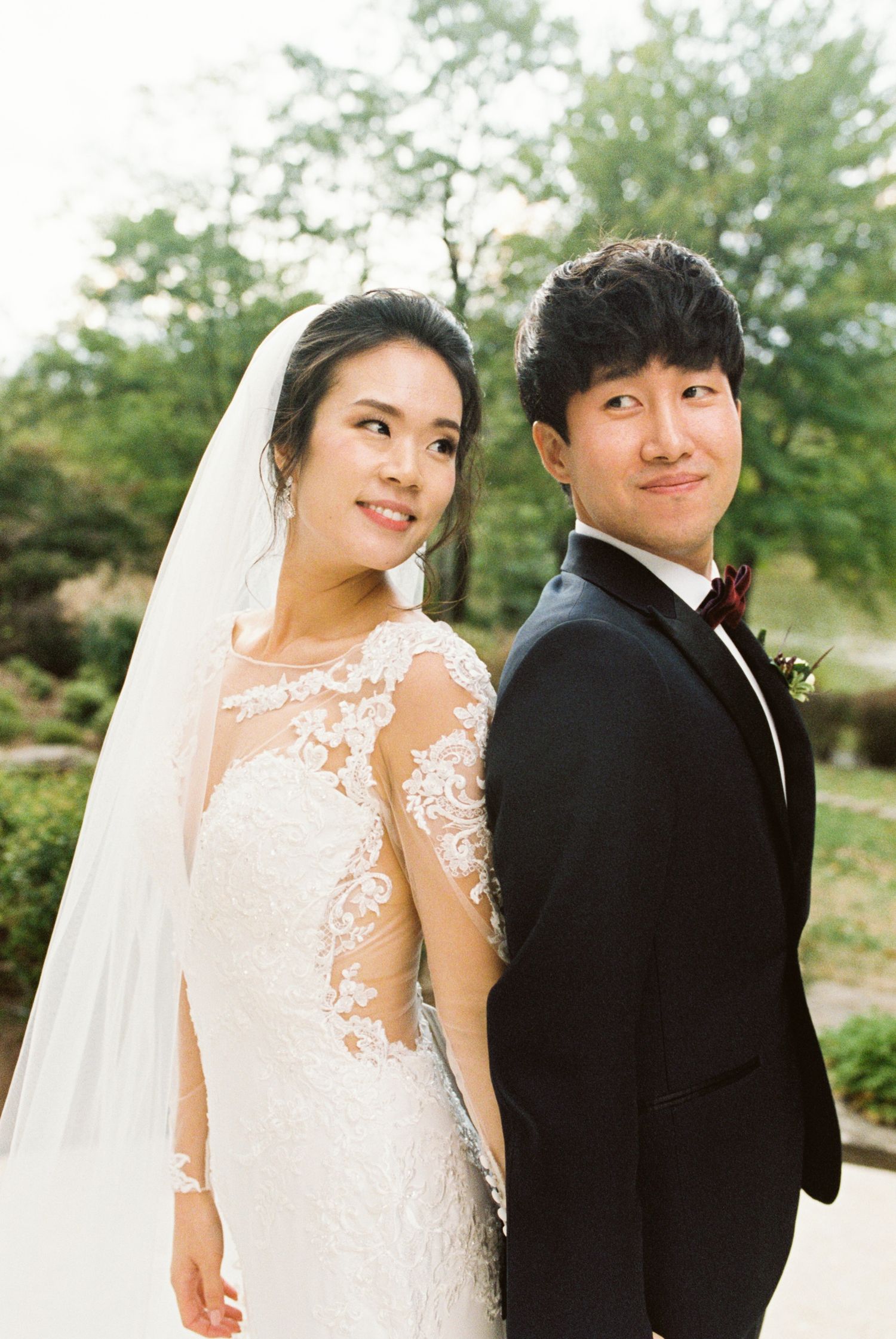 Elegant wedding couple poses outdoors with bride wearing lace dress and flowing veil.