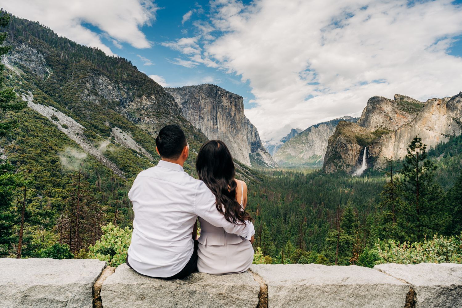 Couple sitting on stone wall at Yosemite Valley overlook with El Capitan and waterfall views.