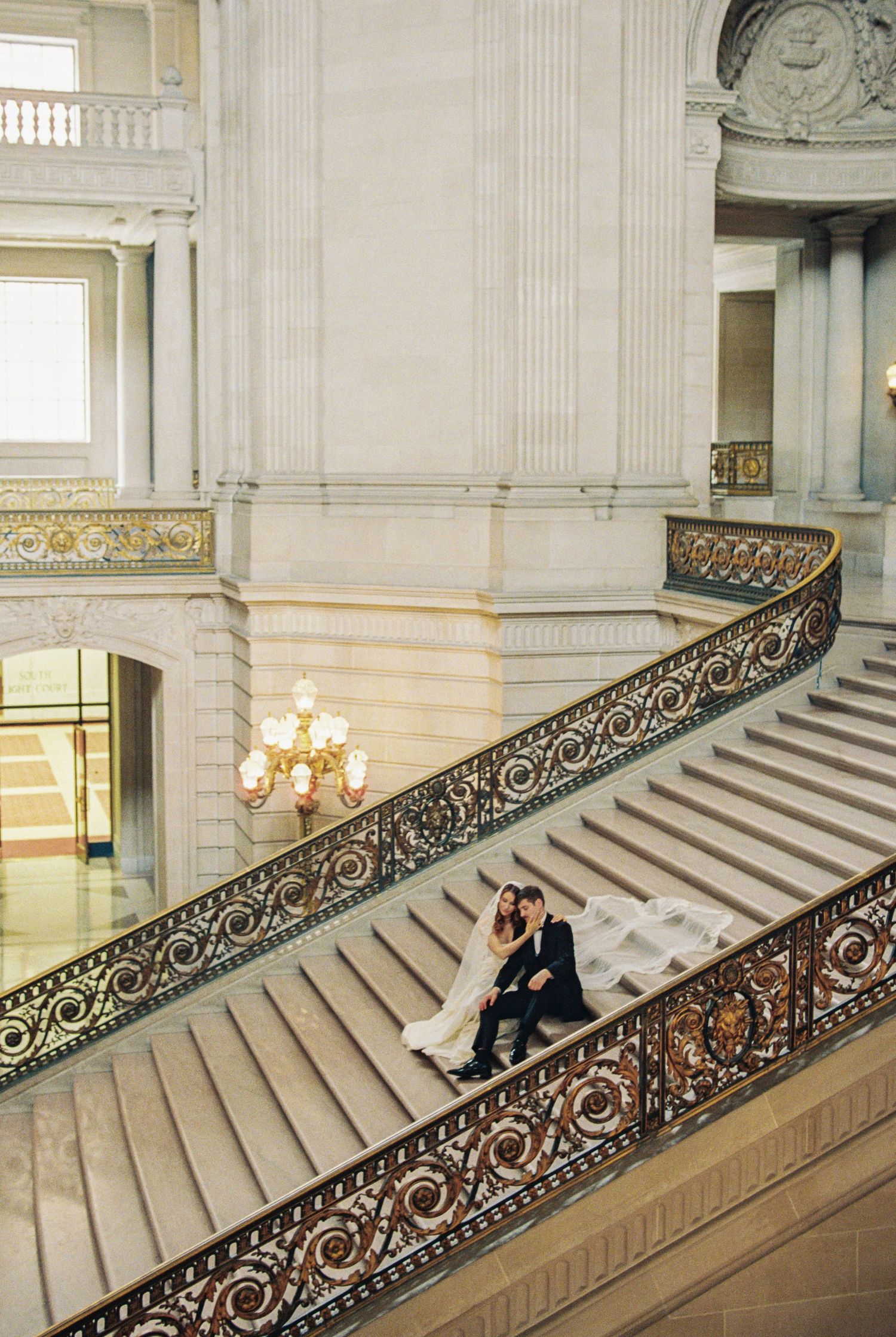 Bride and groom sitting together on ornate grand staircase inside San Francisco City Hall on their wedding day.
