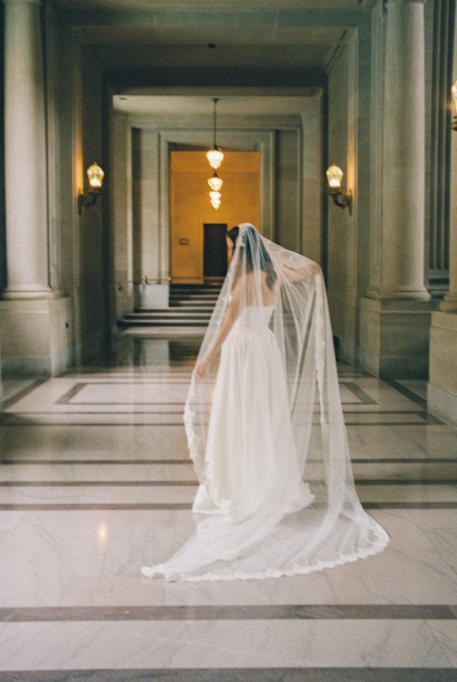 Bride in white gown and long cathedral veil stands in elegant marble hallway with tall columns and warm lighting.