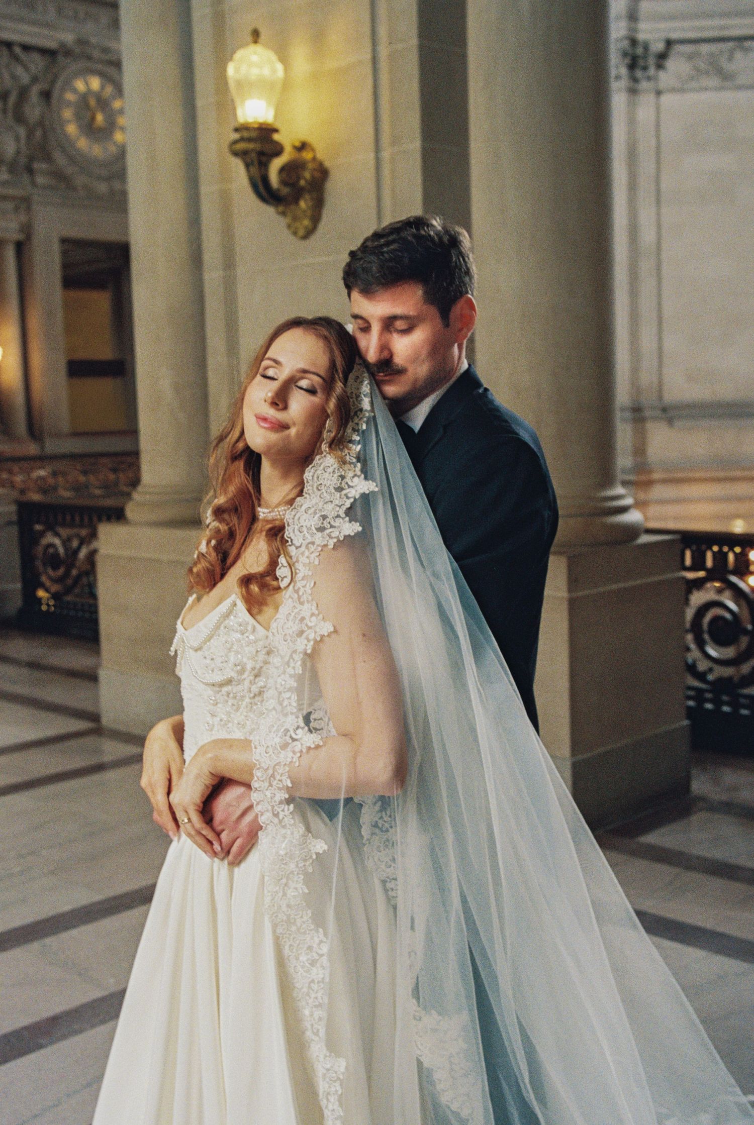 Bride and groom embrace at San Francisco City Hall wedding, bride in lace gown with blue veil, groom in dark suit.